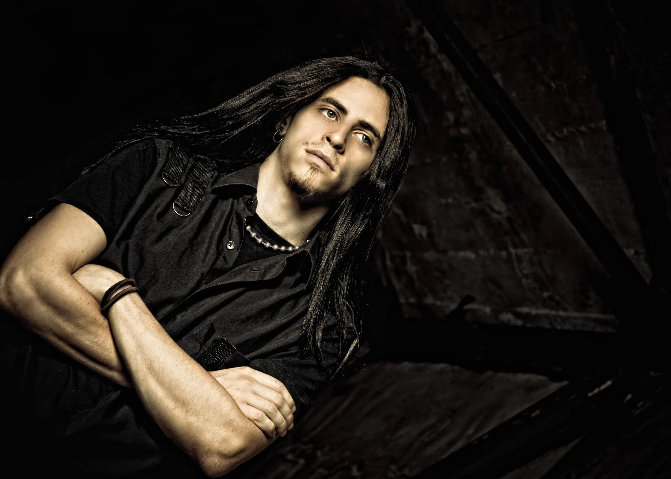 Man with long black hair in black metal attire posing against dark industrial background. 
