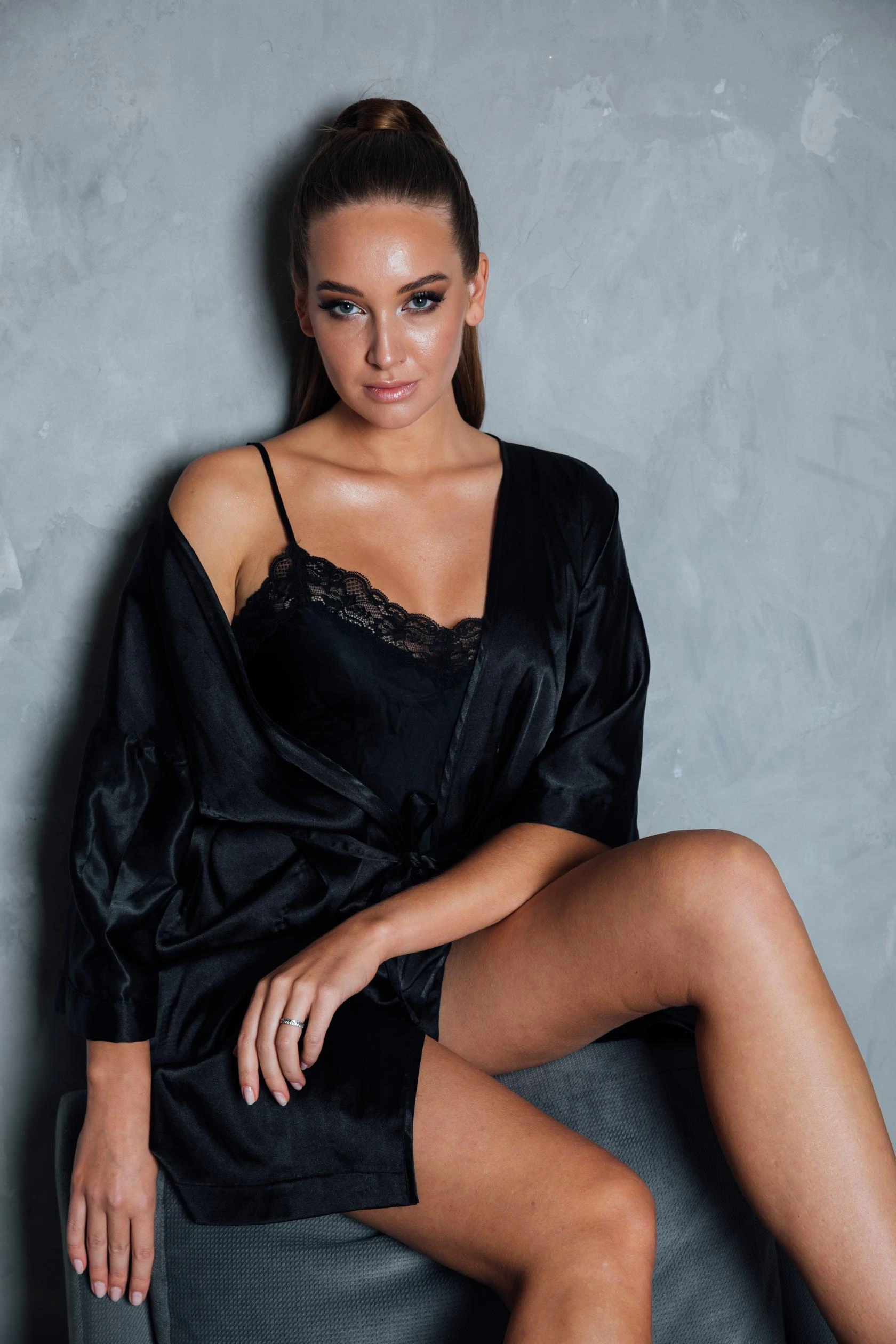 Woman in a black satin nightgown and robe sitting against a textured grey wall.
