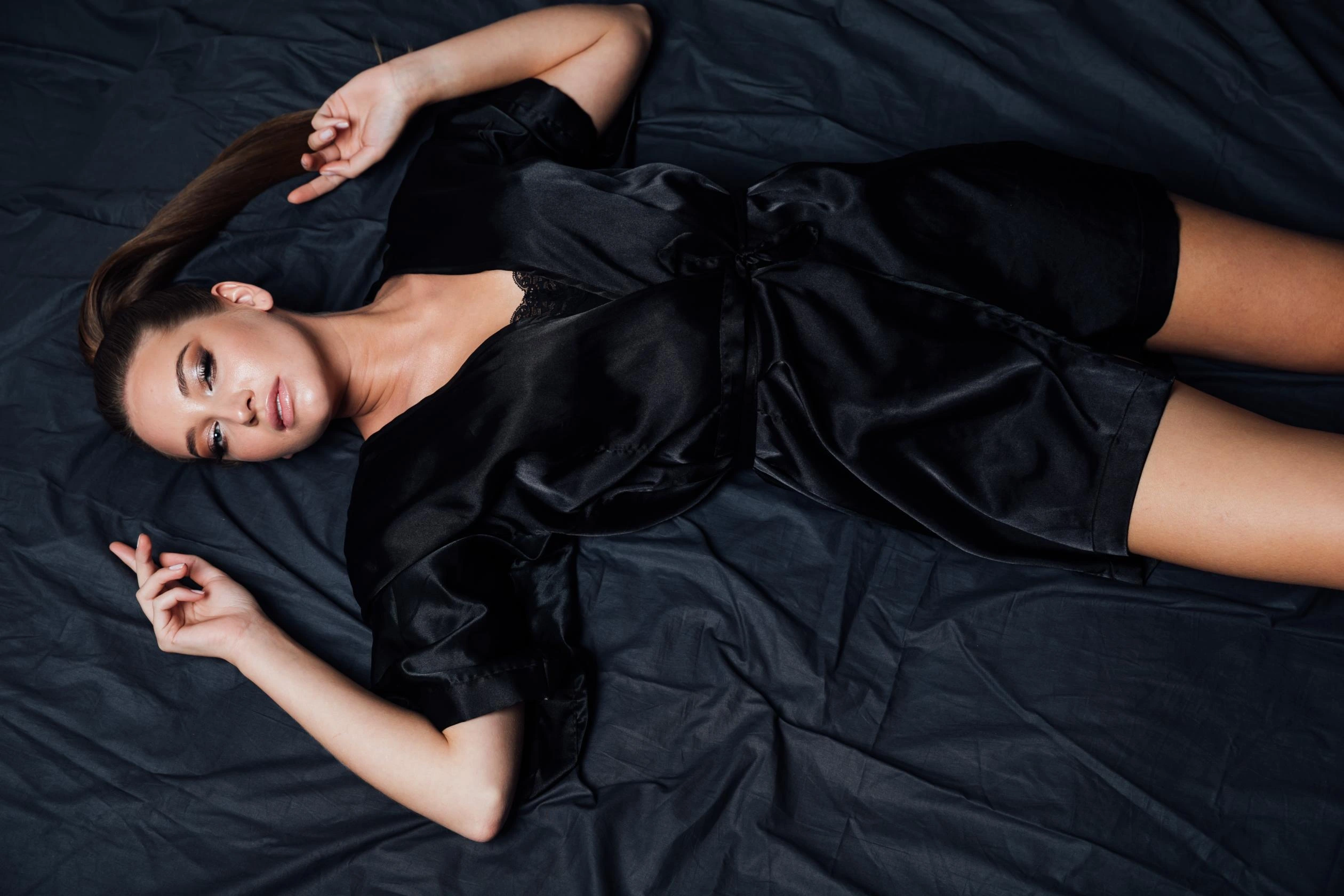 Woman lying on a dark bed in a black satin robe, gazing into the distance.