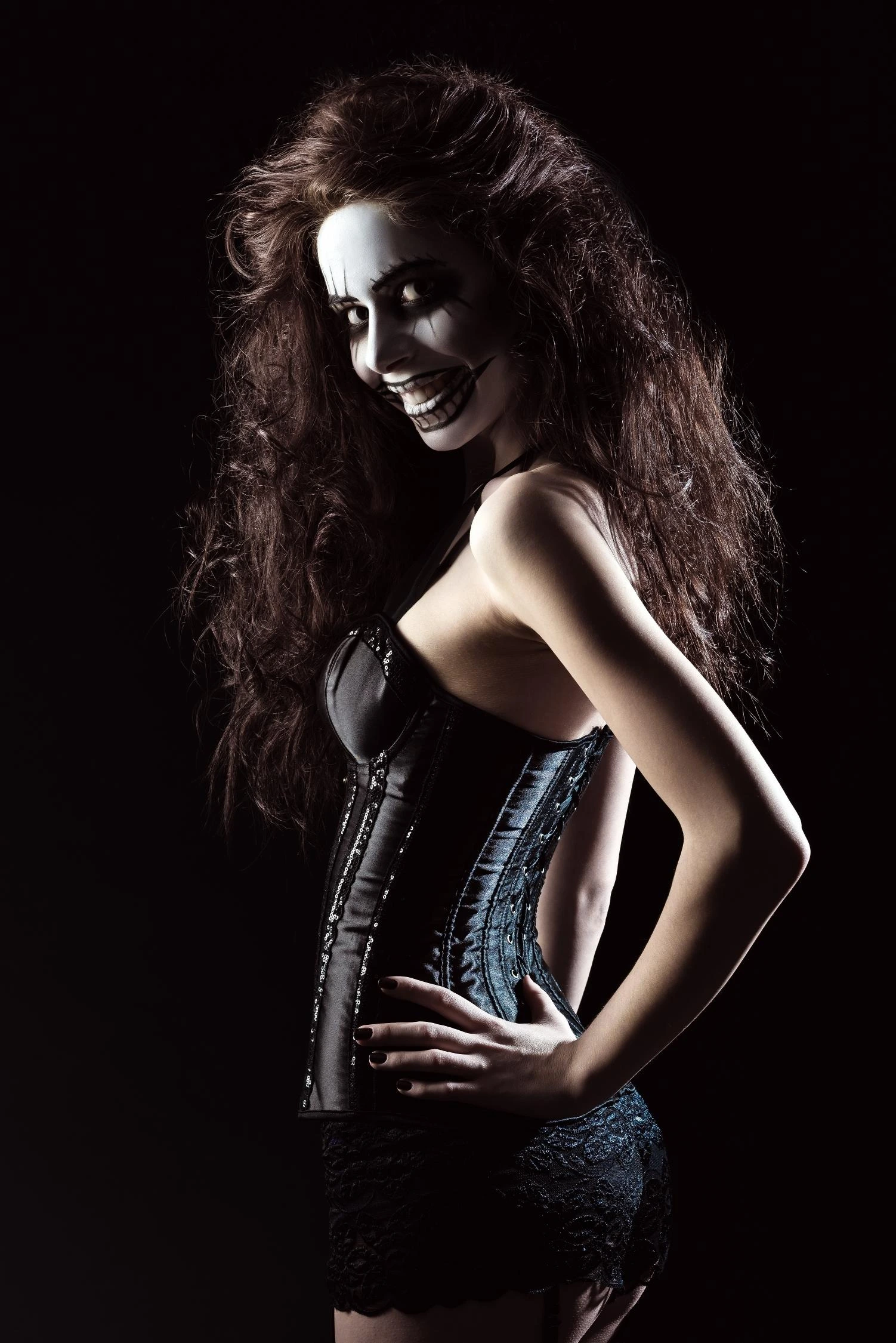 Woman in a black corset and mini skirt with Halloween-style skull makeup, smiling in a dark studio.