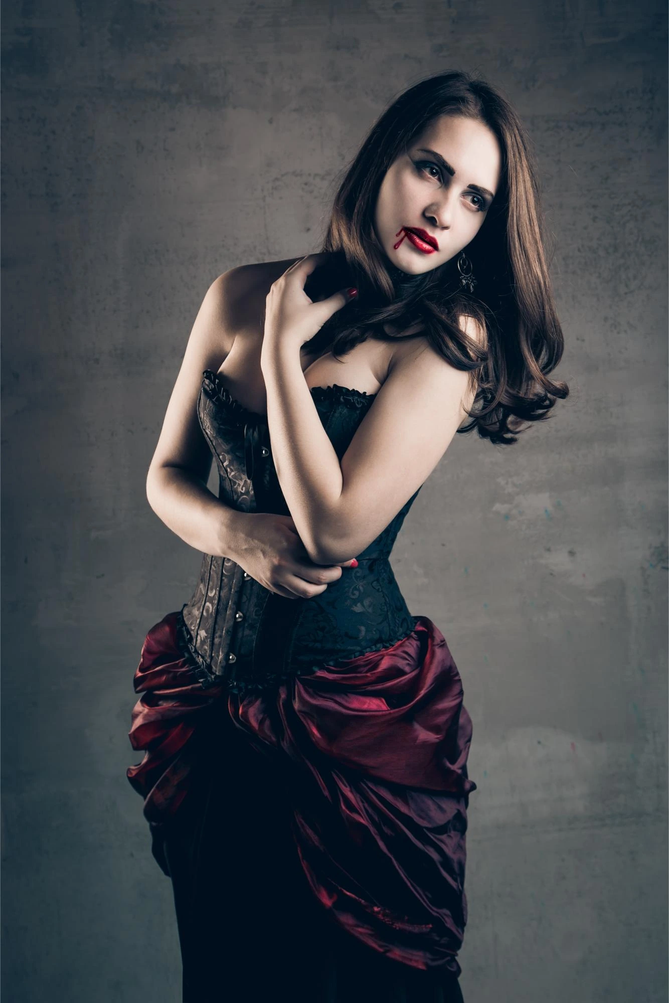 Gothic woman in black corset and red skirt with vampire makeup and blood on lips, standing against a dark background.