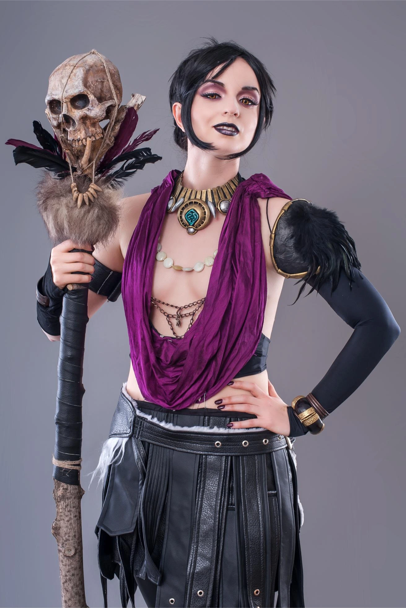Woman in fantasy tribal goth costume with skull staff and dark makeup, wearing purple and black with confident expression.