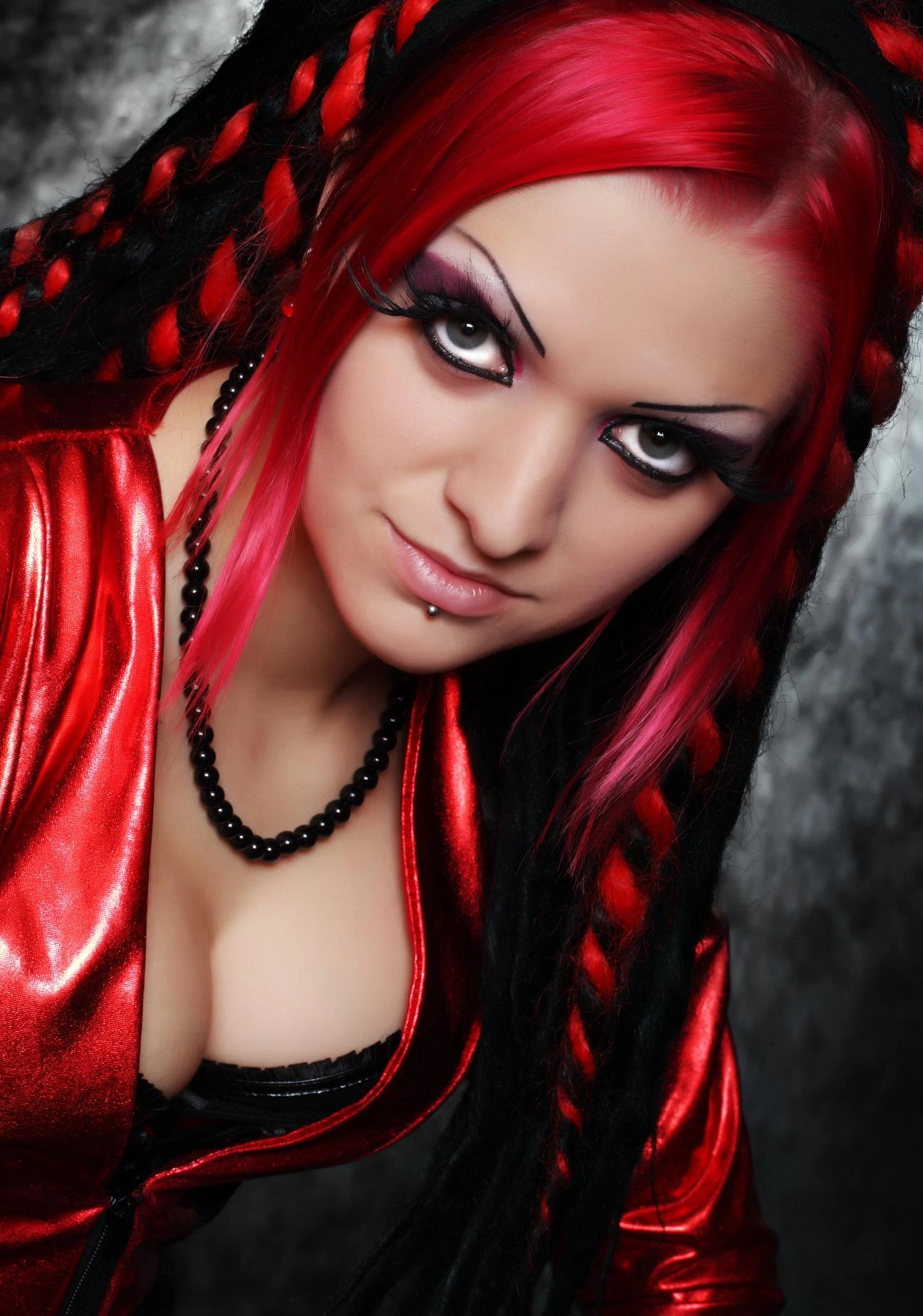 Close-up portrait of a gothic woman with striking red and black braids, intense eye makeup, and a deep neckline in a red PVC jacket.