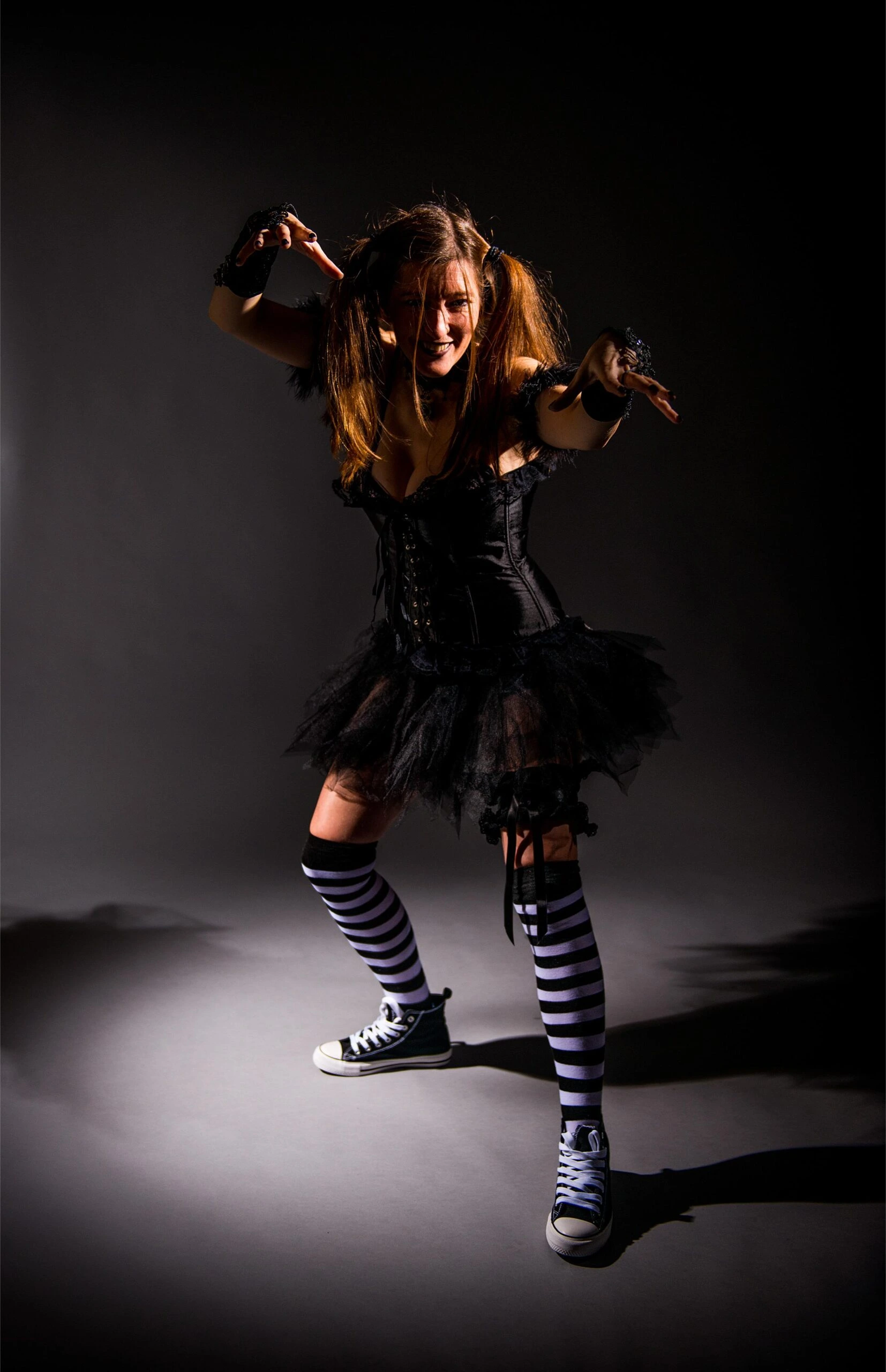 Dark punk goth girl in a black tutu-style mini skirt and corset, wearing striped stockings and Converse sneakers, posing in a dramatic spotlight.