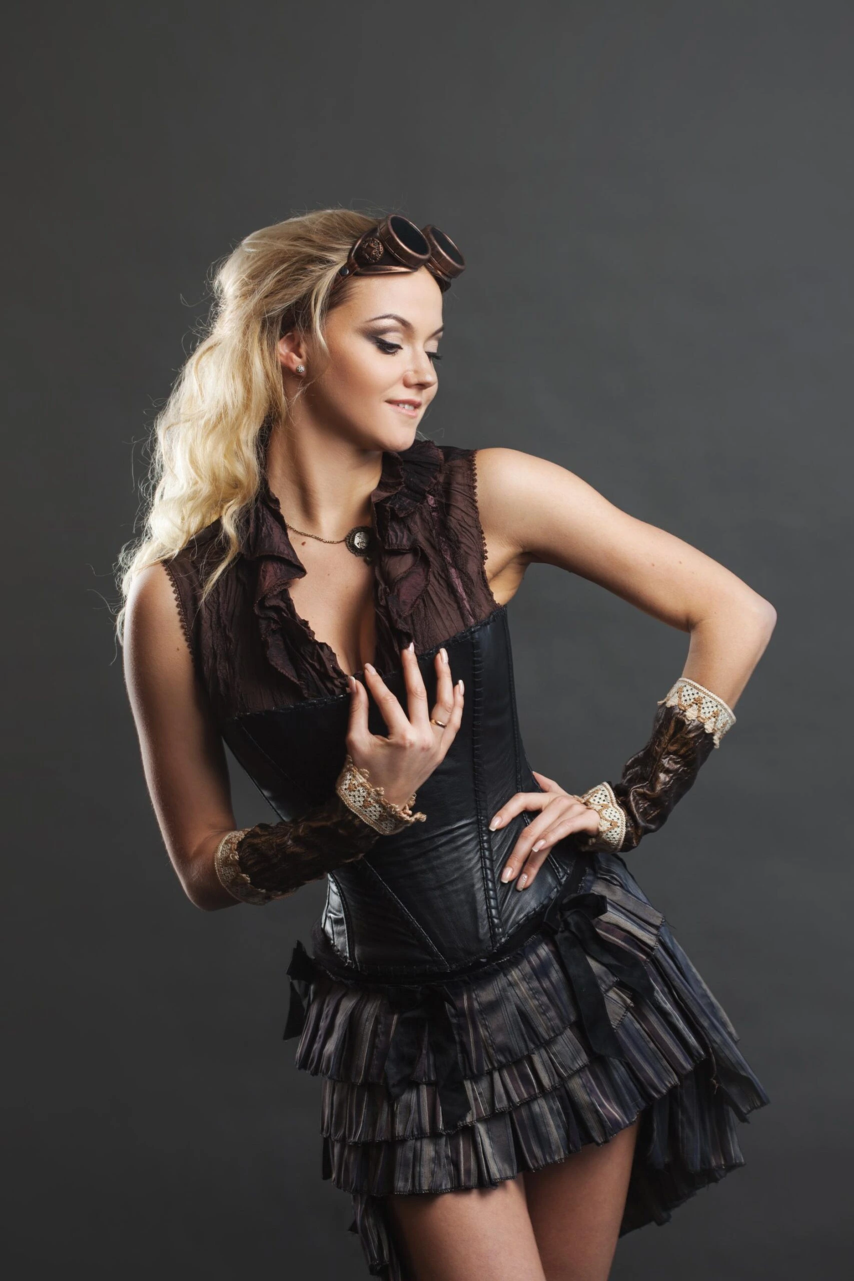 Blonde woman in a black corset and layered ruffled mini skirt with lace-trimmed fingerless gloves, styled with steampunk goggles on her head.