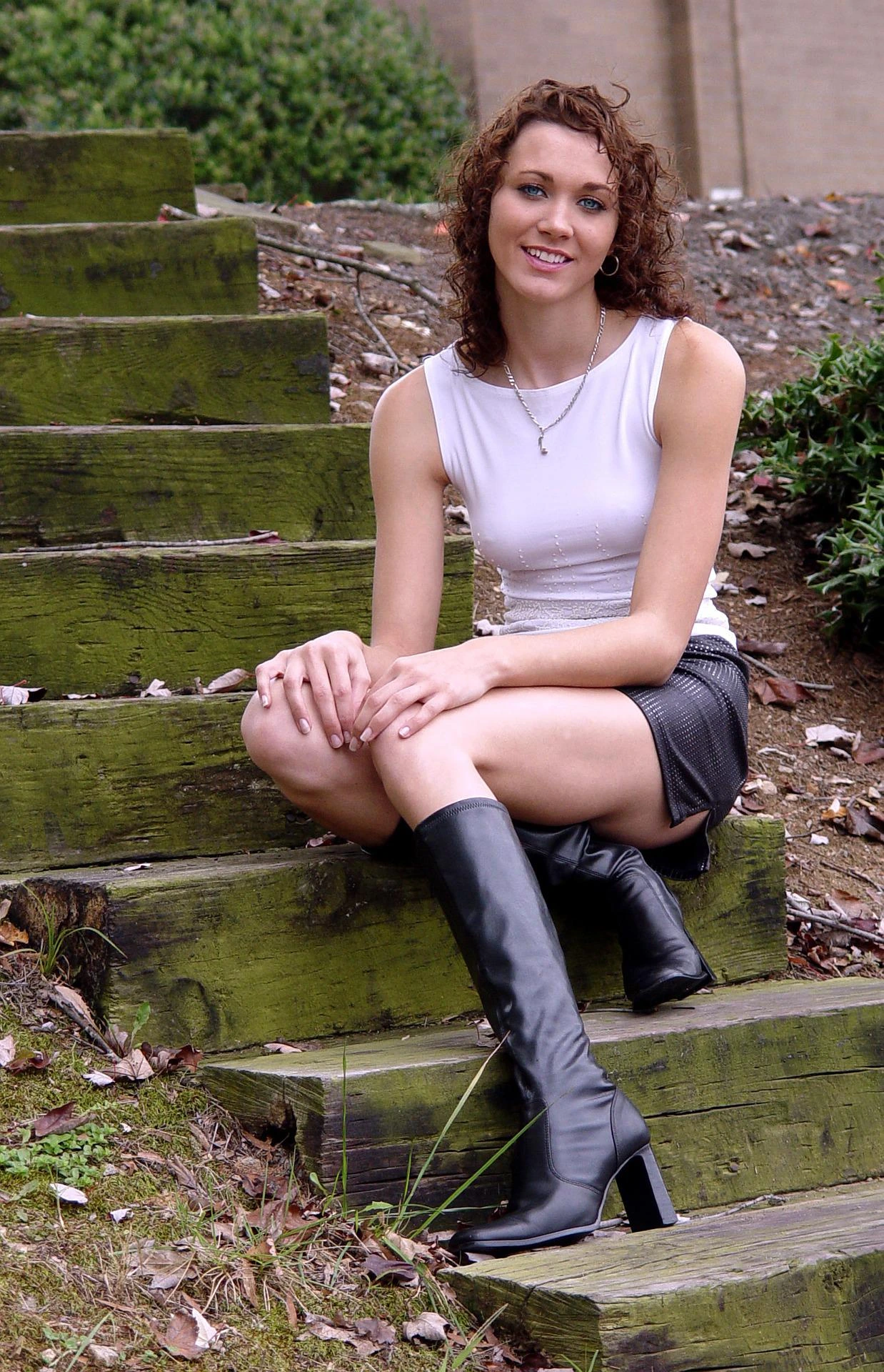 Woman sitting on mossy wooden steps wearing a black leather mini skirt and knee-high boots