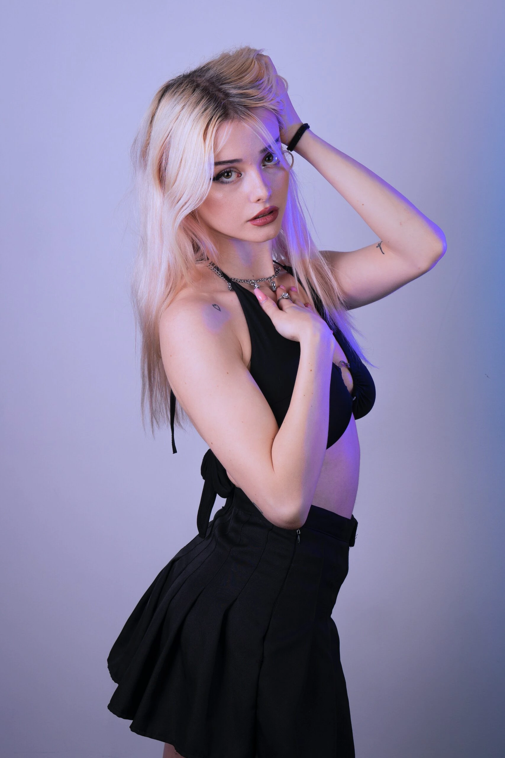 Blonde woman in black pleated mini skirt and halter top posing under purple lighting