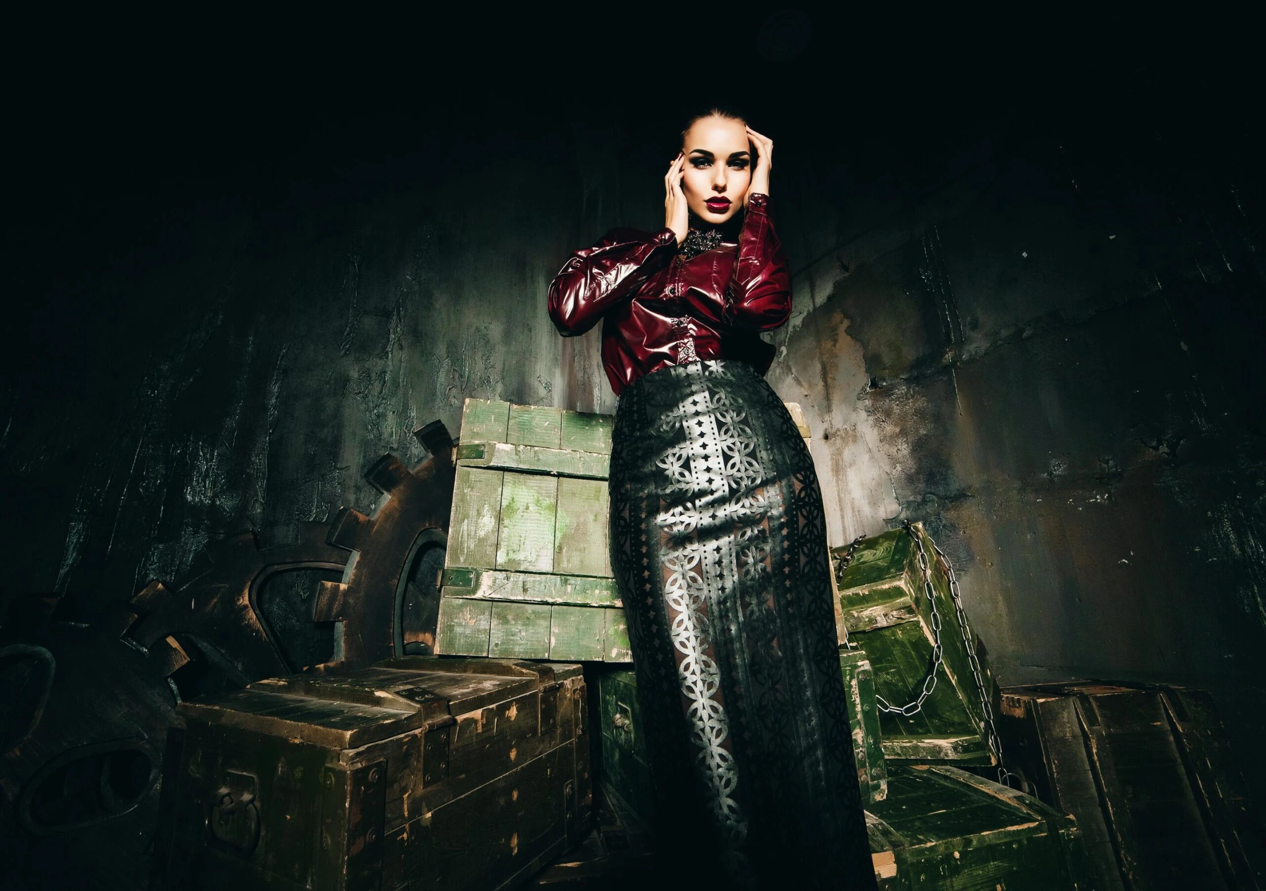 Woman in burgundy latex top and black leather pencil skirt standing against industrial crates