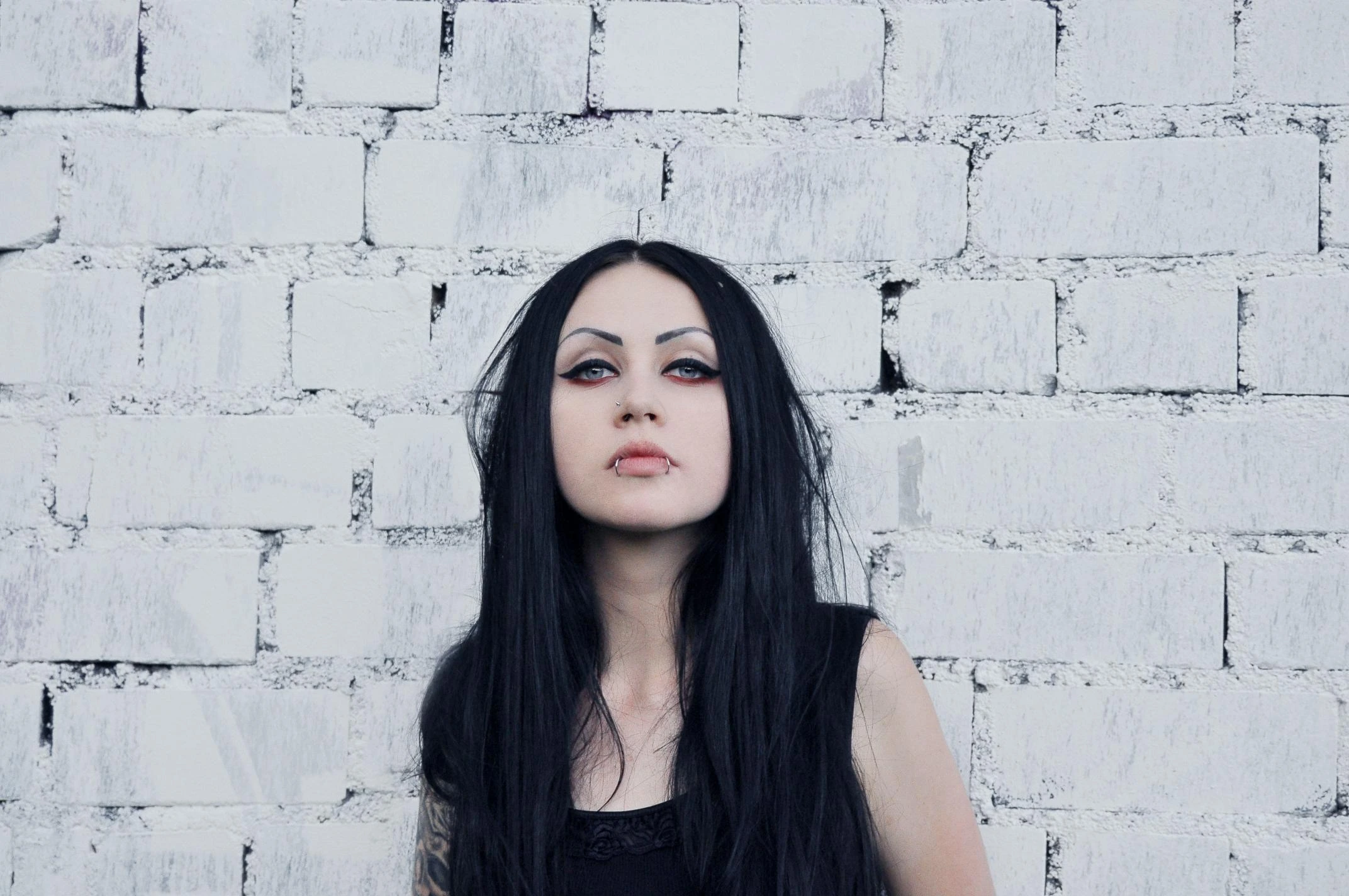 Alternative woman with long black hair and red eyeliner standing against a white-painted brick wall, piercing lip jewelry visible.