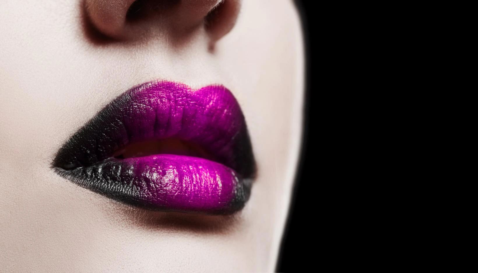 Close-up of glossy ombré lips with deep black fading into vibrant purple on porcelain skin, against a black background.