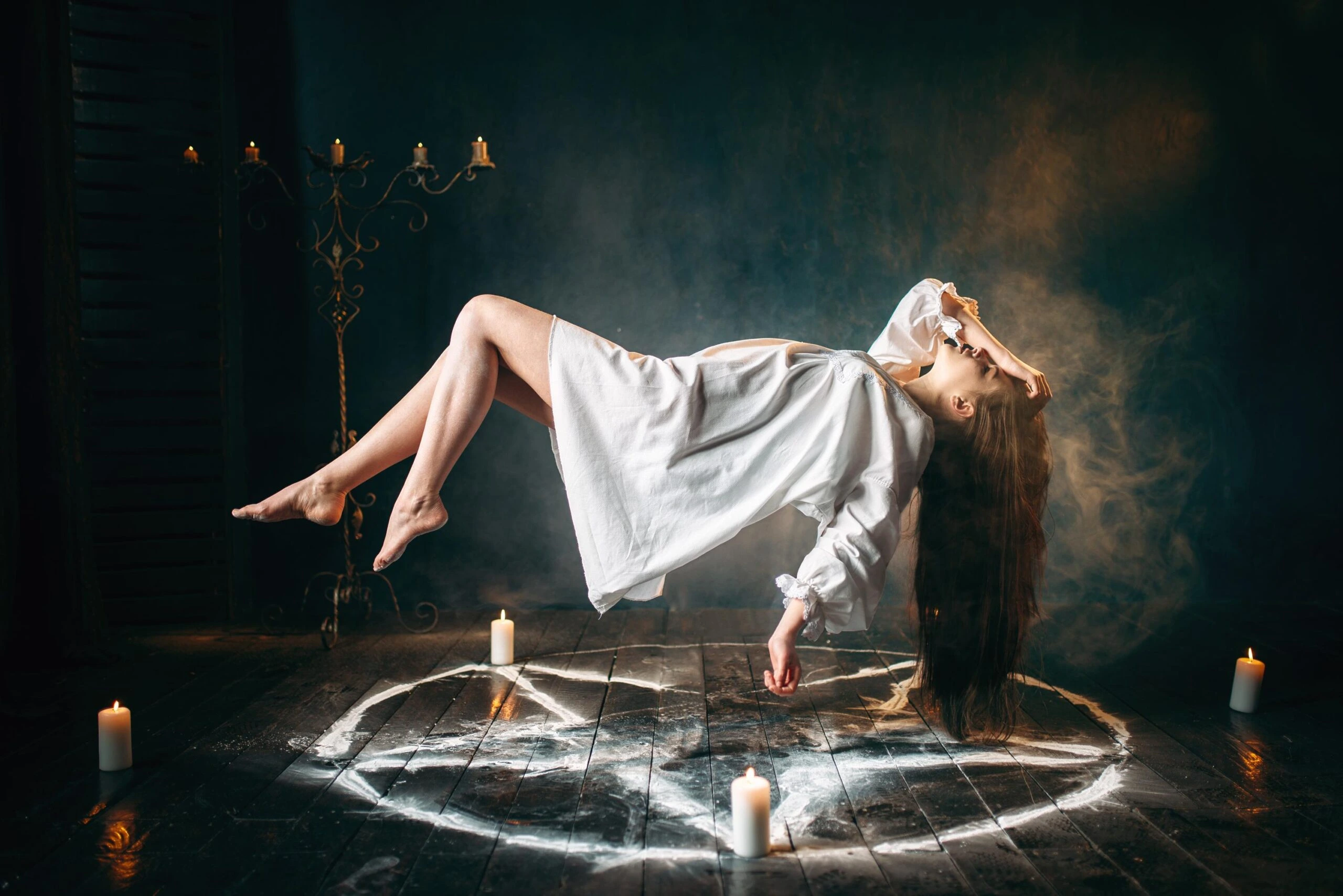 Woman in white nightgown levitating above a pentagram surrounded by candles in a dark ritual chamber.