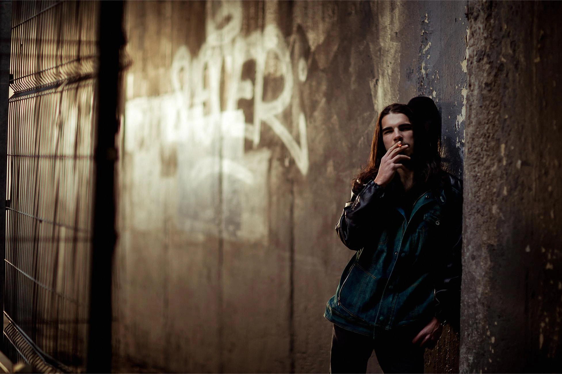 Man with long hair smoking in a gritty urban alley, lit by golden light and graffiti-stained walls.