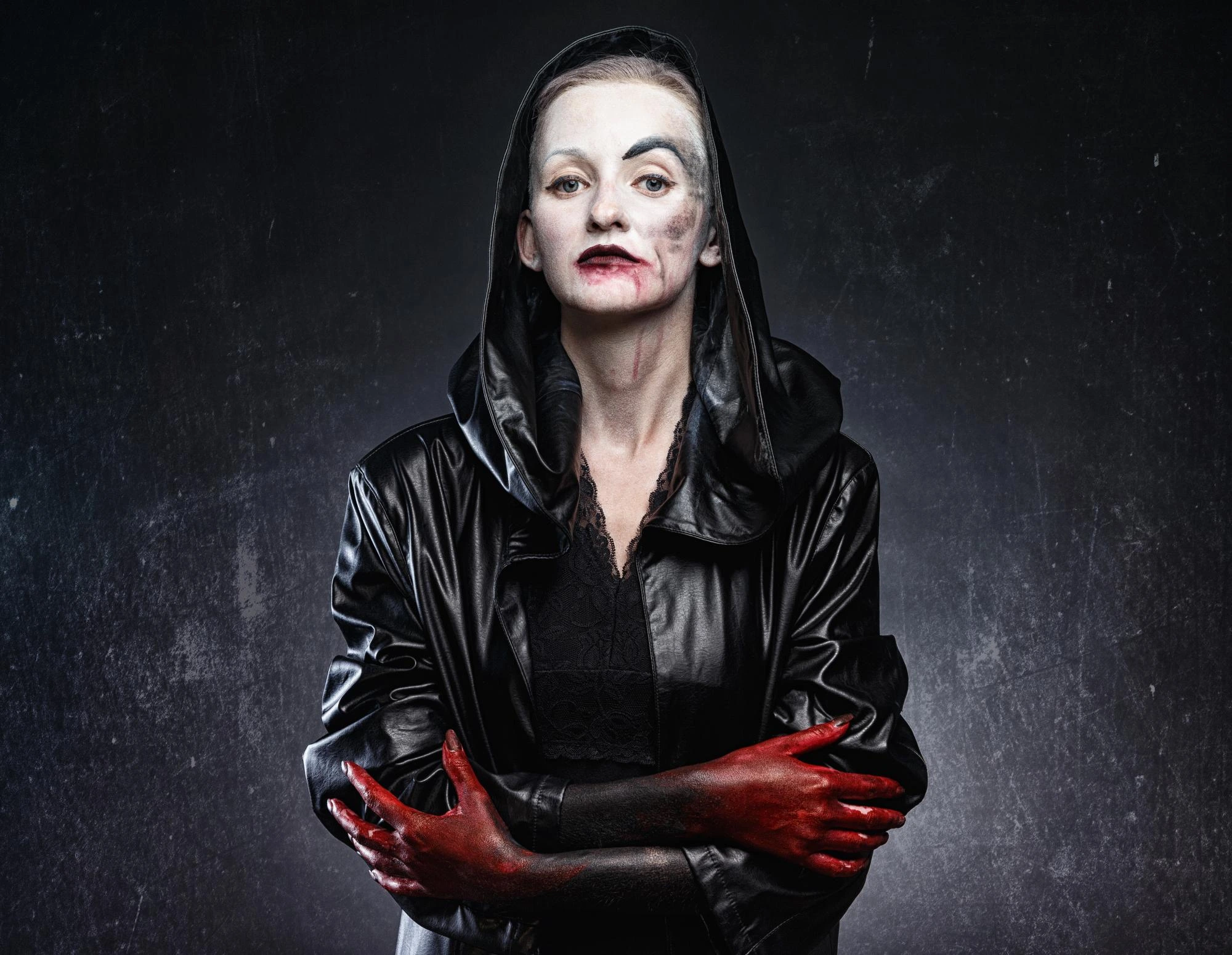 Pale woman in a black hooded coat with blood-smeared hands and gothic makeup, staring directly into camera.