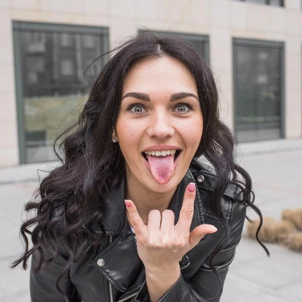 Playful gothic woman in leather jacket sticking out tongue and making rock hand sign outdoors.