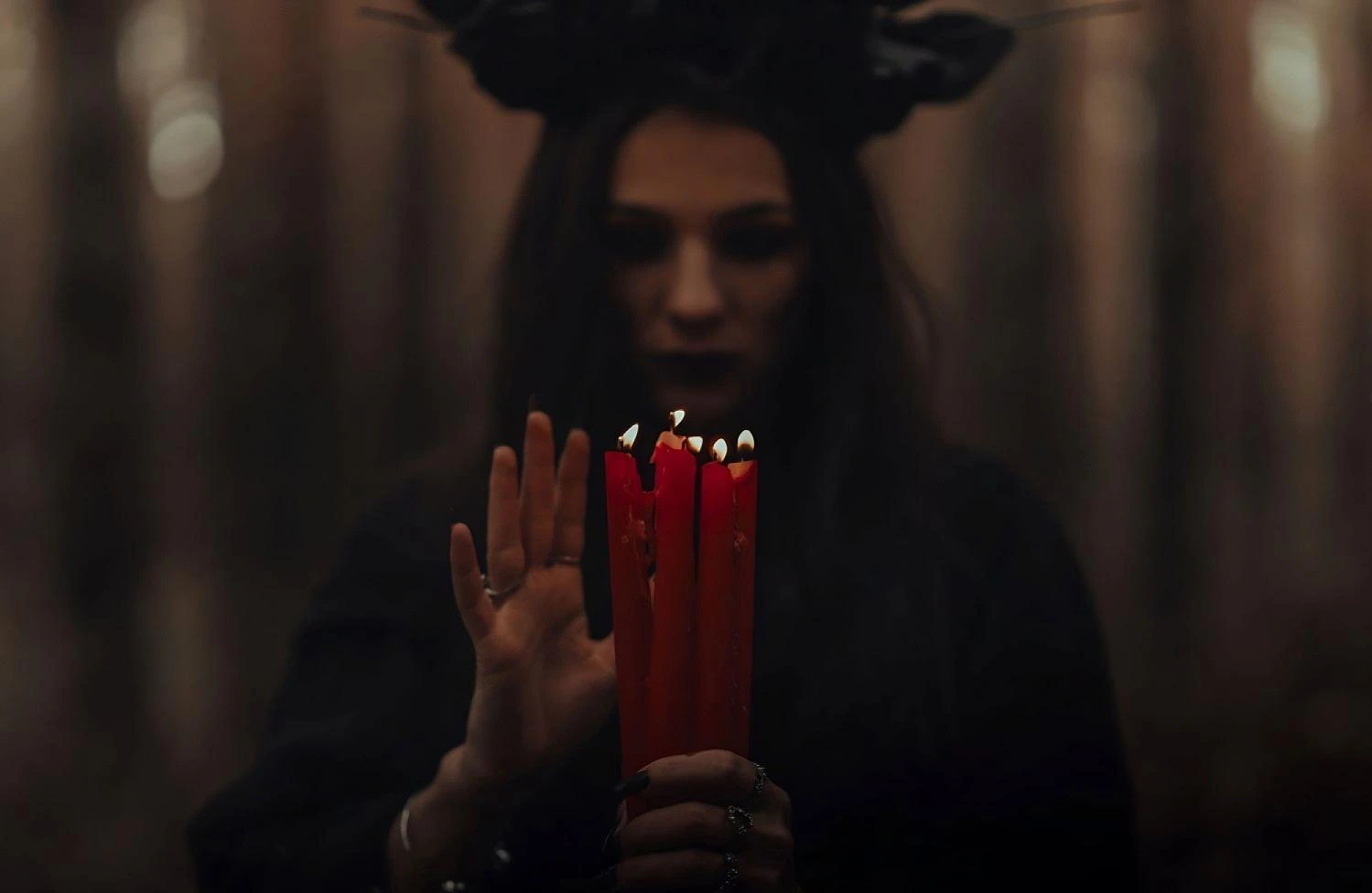 Mysterious goth woman with dark floral crown, holding burning red candles in a misty forest at dusk.