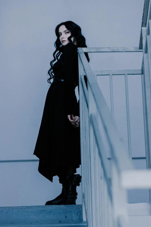Woman in gothic outfit standing on stairwell, turning back with fierce gaze in a moody, blue-lit scene.
