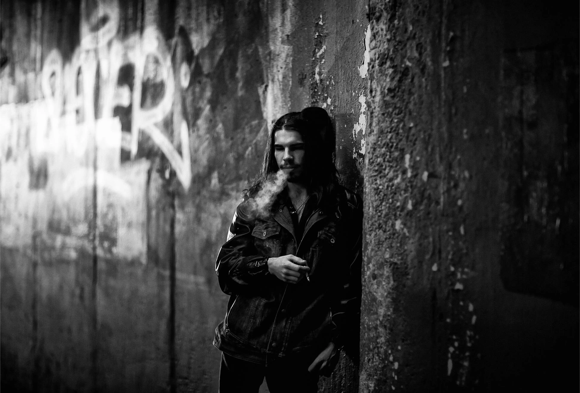 Man with long hair smoking in a dark alley against a graffiti-covered wall