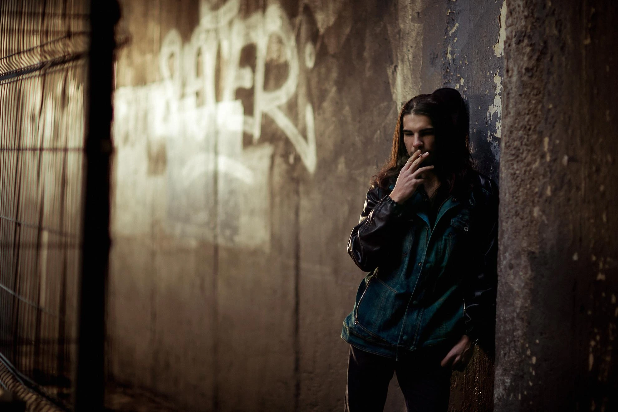 Man with long hair smokes a cigarette against a graffiti wall in a dim urban tunnel.