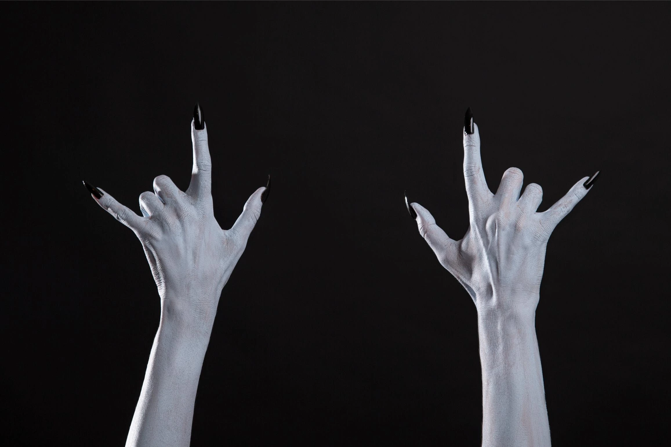 Two pale hands with long black nails raised in devil horns against black background