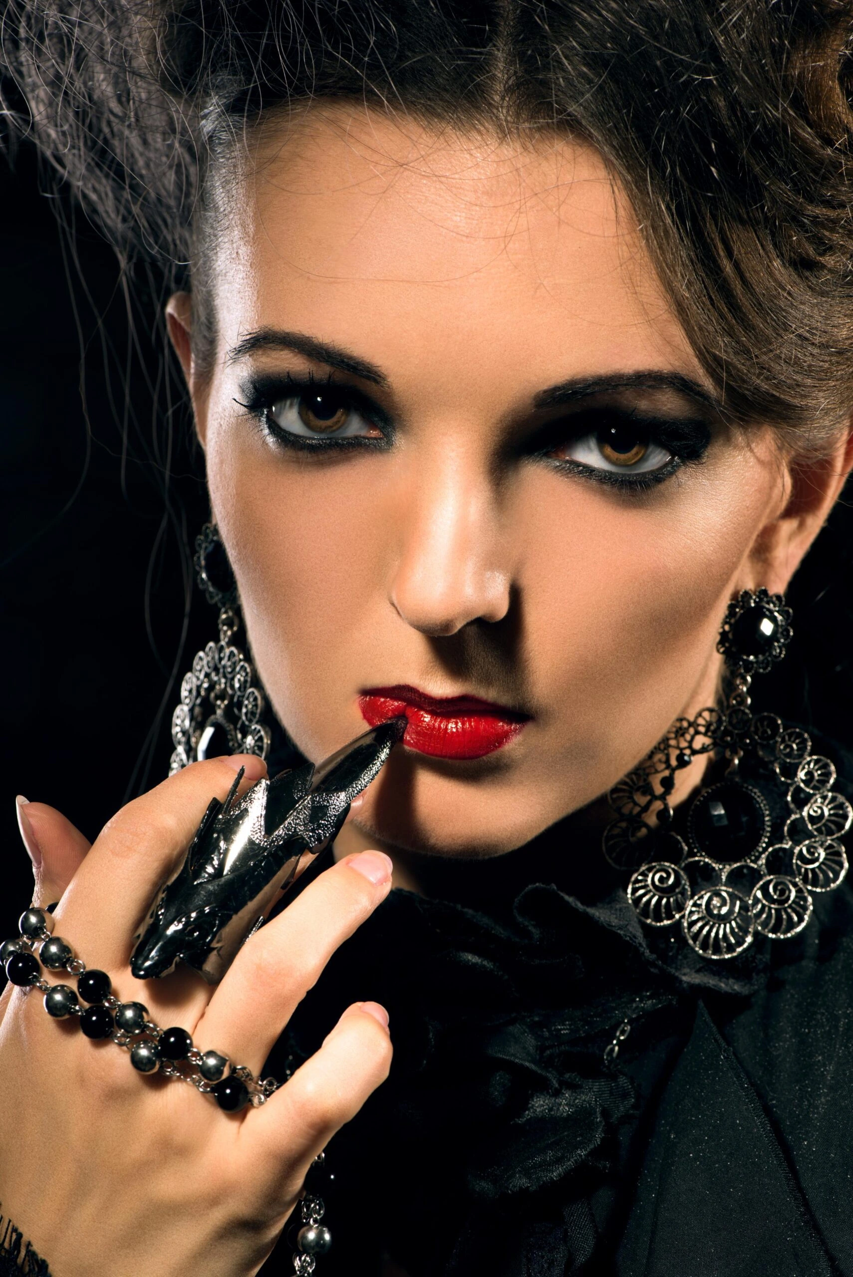 Close-up of woman with red lips, gothic jewelry and dramatic black eyeliner holding finger to mouth