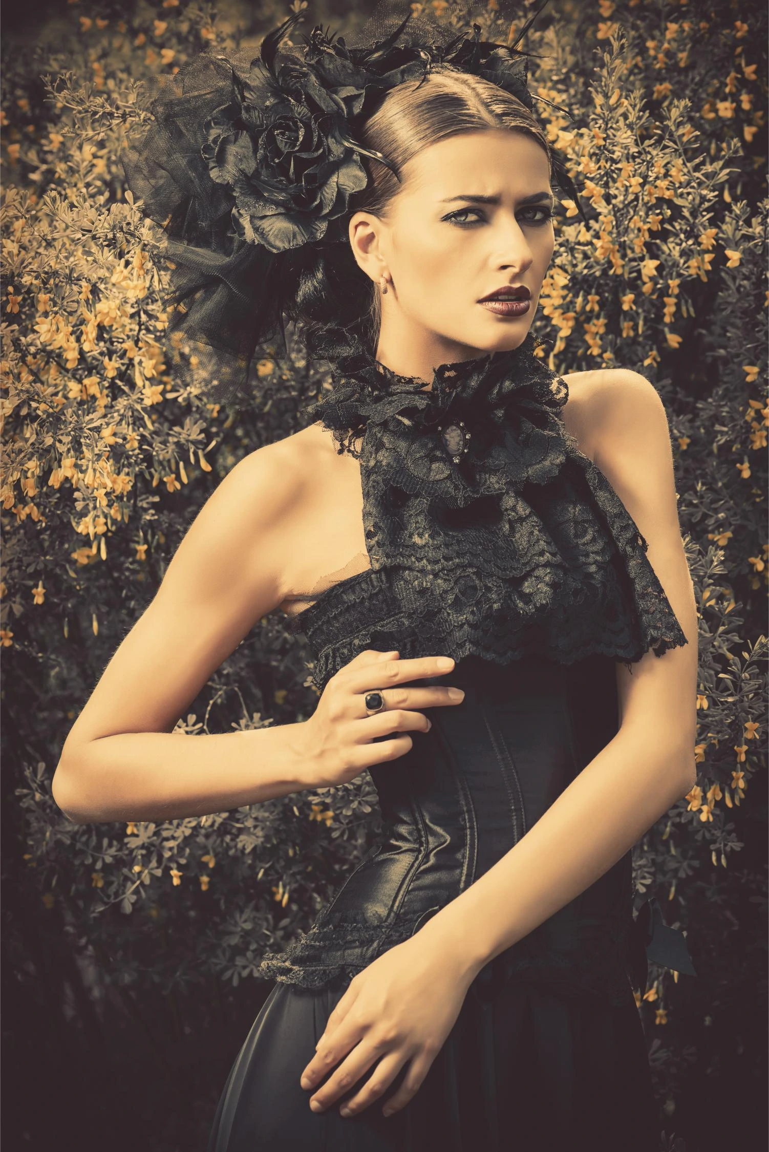 Gothic woman in black lace corset and large floral headpiece posing against yellow flowering bushes.