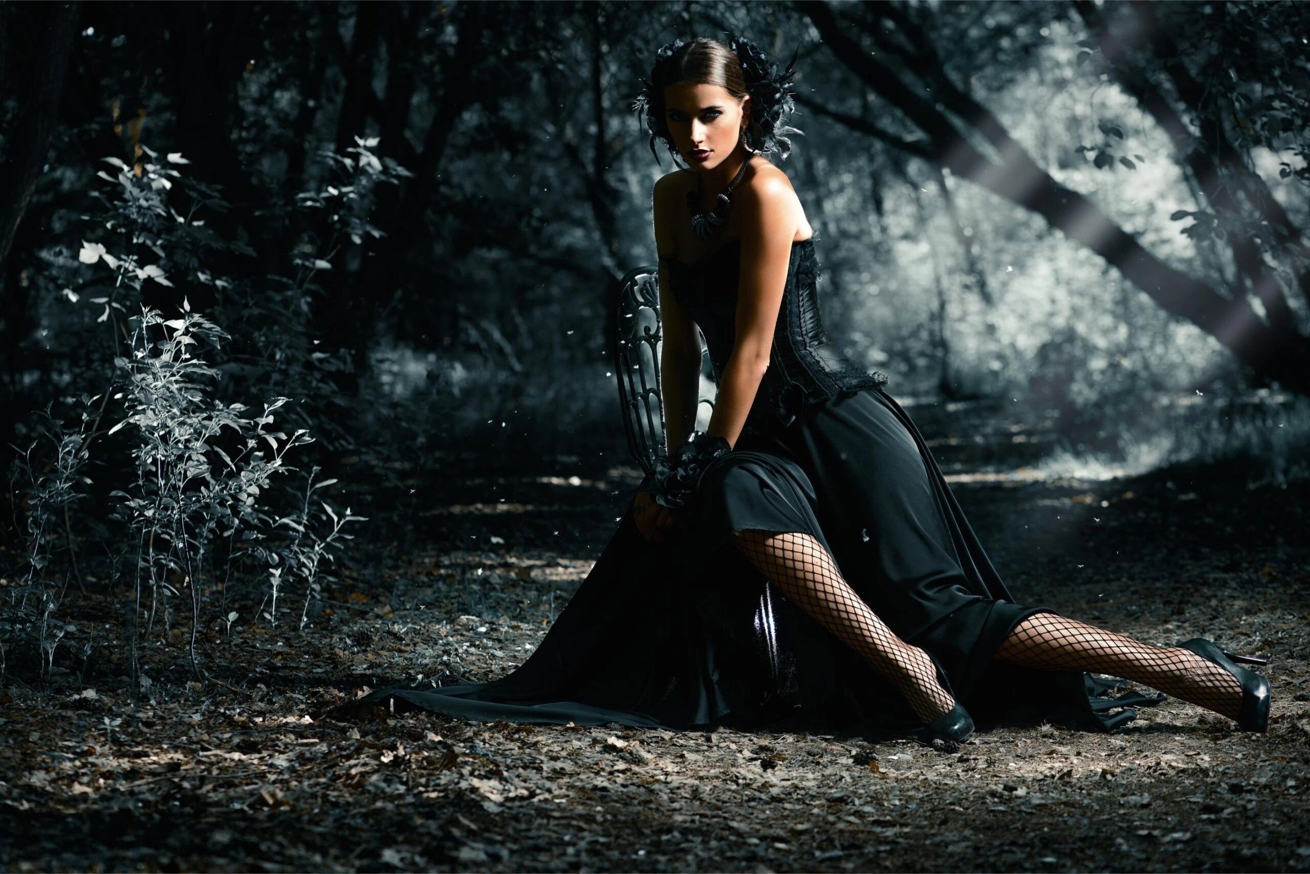 Gothic woman in black corset and fishnet tights kneeling in a dark forest, dramatic lighting.