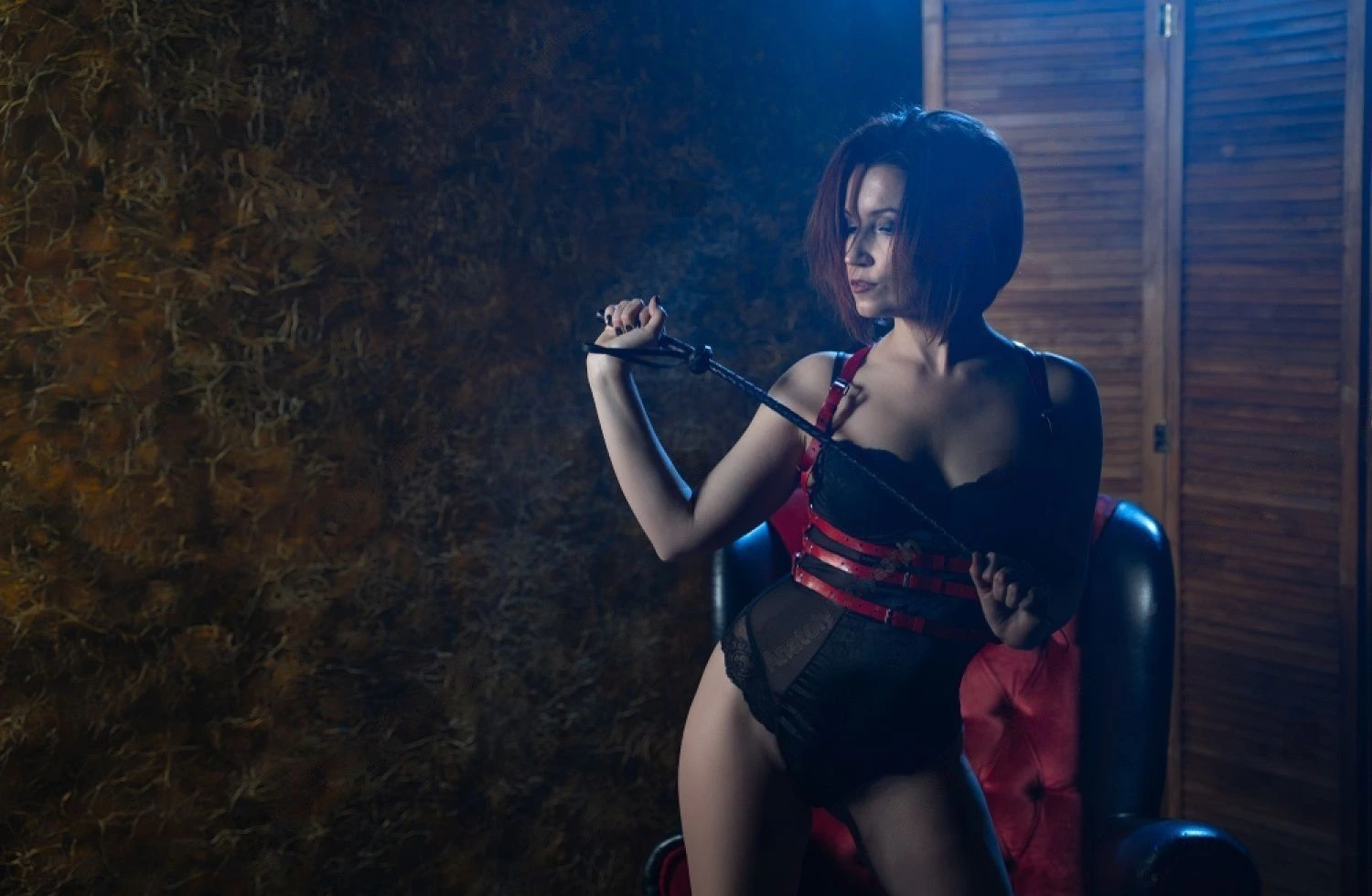  Woman with short red hair in black and red bondage gear holding a whip, posing with dominant attitude in cool blue stage light.
