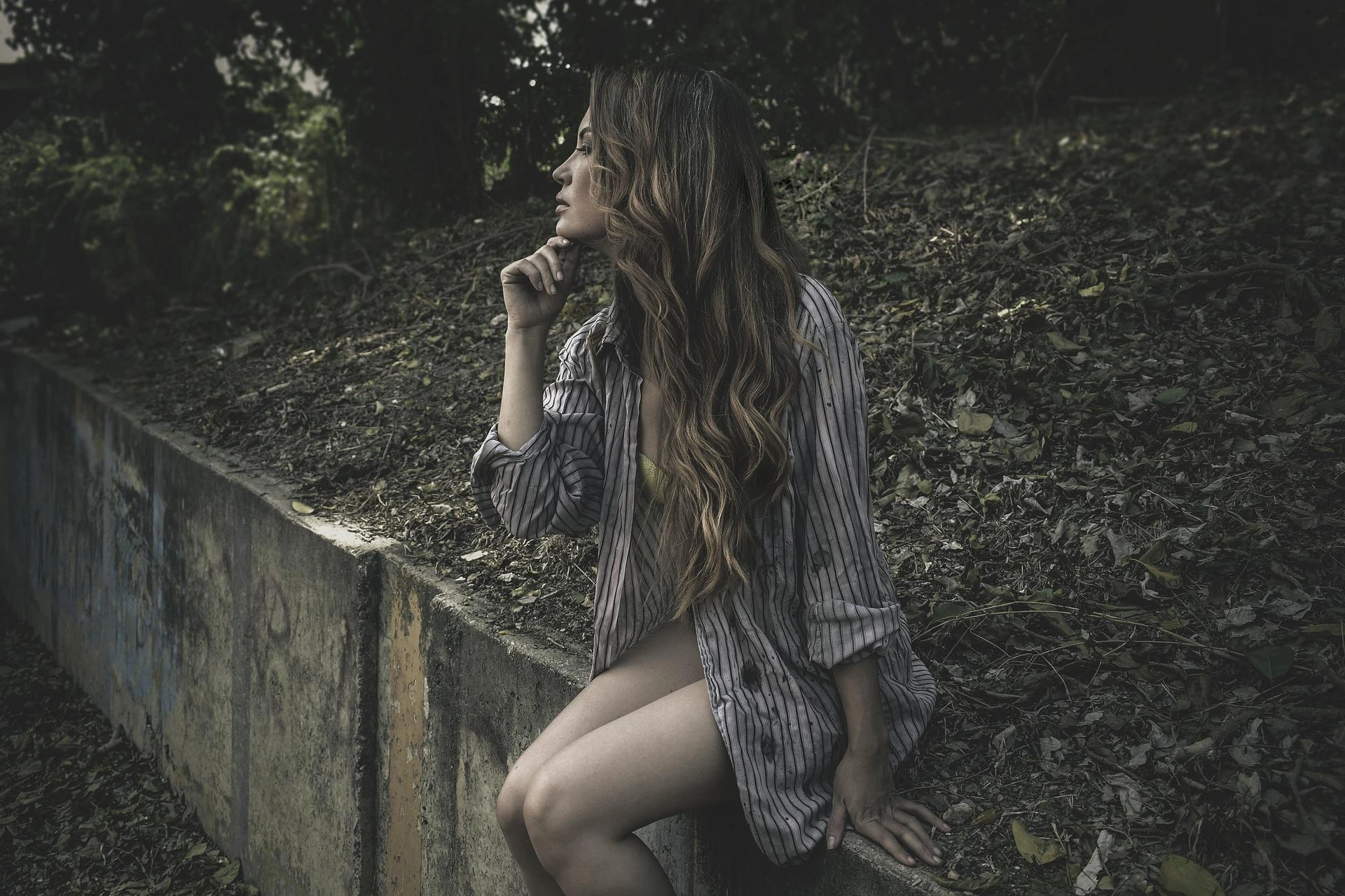 Woman in an oversized shirt sitting by a concrete ledge surrounded by leaves, with a dreamy yet dark natural expression.