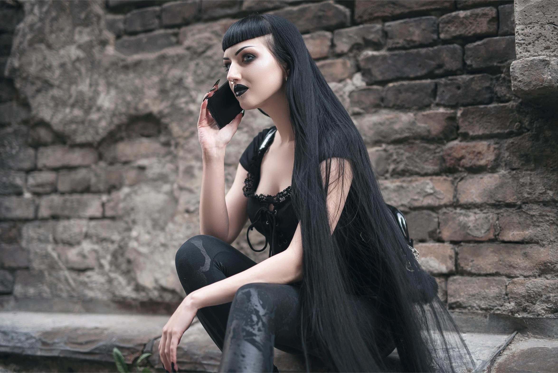 Goth woman in black clothes talking on a smartphone against a brick wall