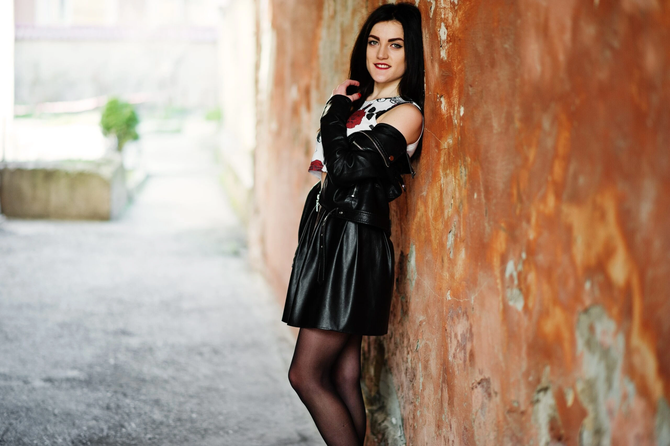 A person leaning against a textured brown wall, wearing a black leather jacket, a printed top, and a black pleated mini skirt.
