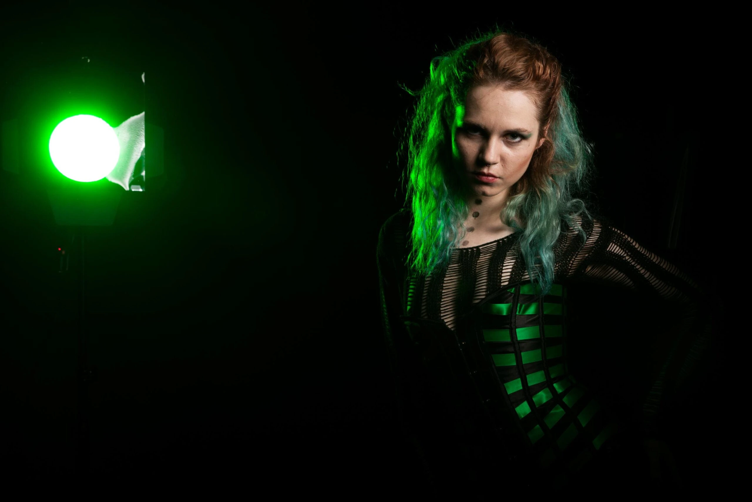 Gothic woman with green-highlighted hair wearing a black corset outfit under dramatic green stage lighting.