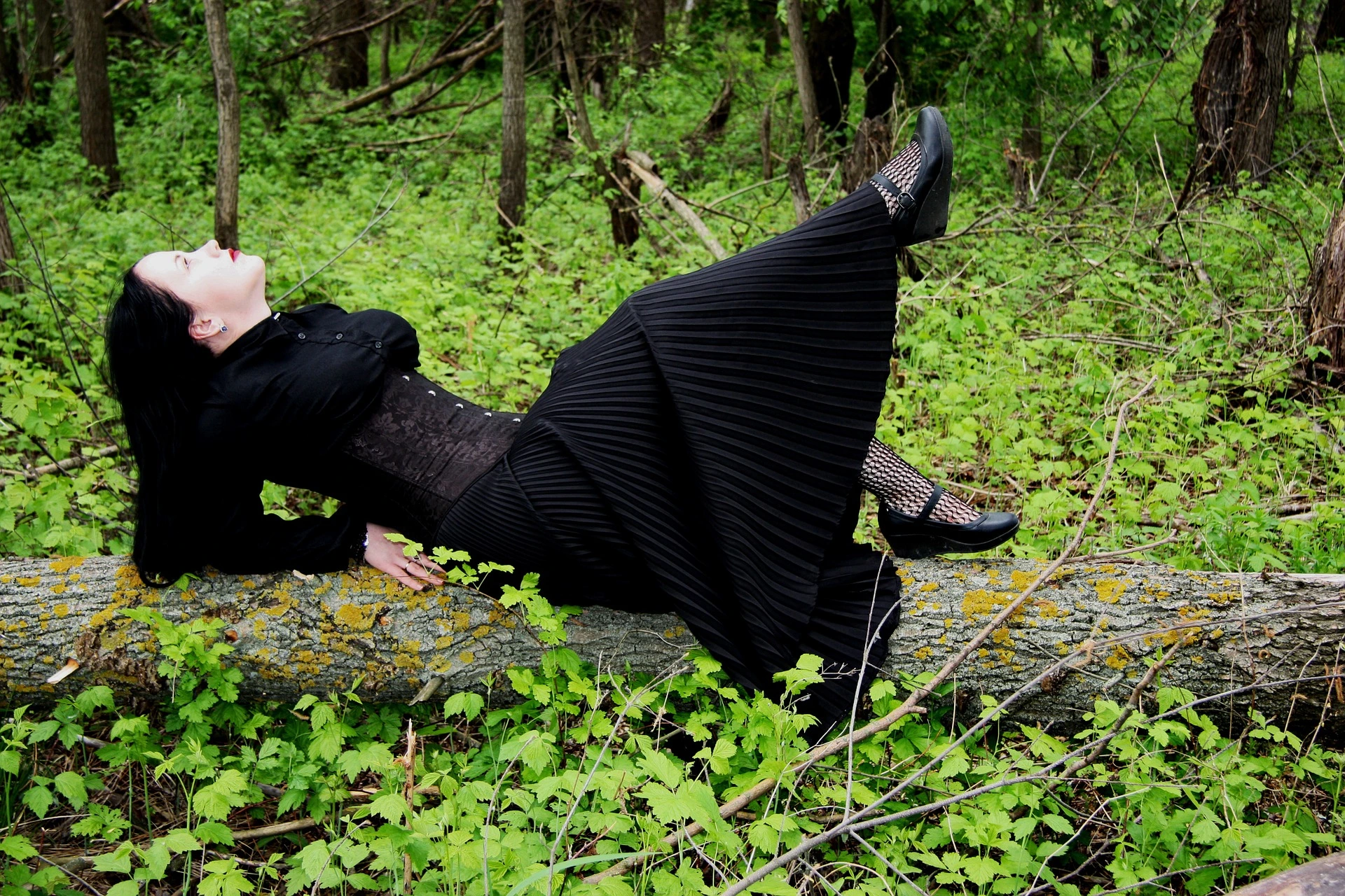Goth woman kneeling in the woods in a black dress, a corset, and white flowers on her arms.