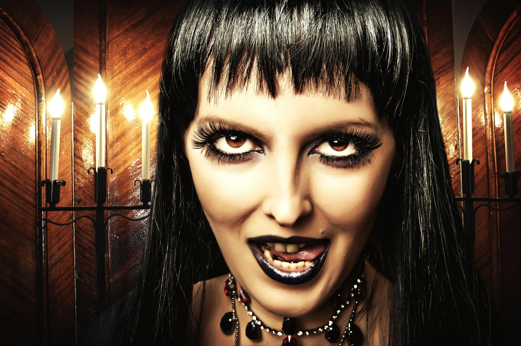 Gothic woman with black hair, dark makeup, and black lipstick smirking in candlelit room.