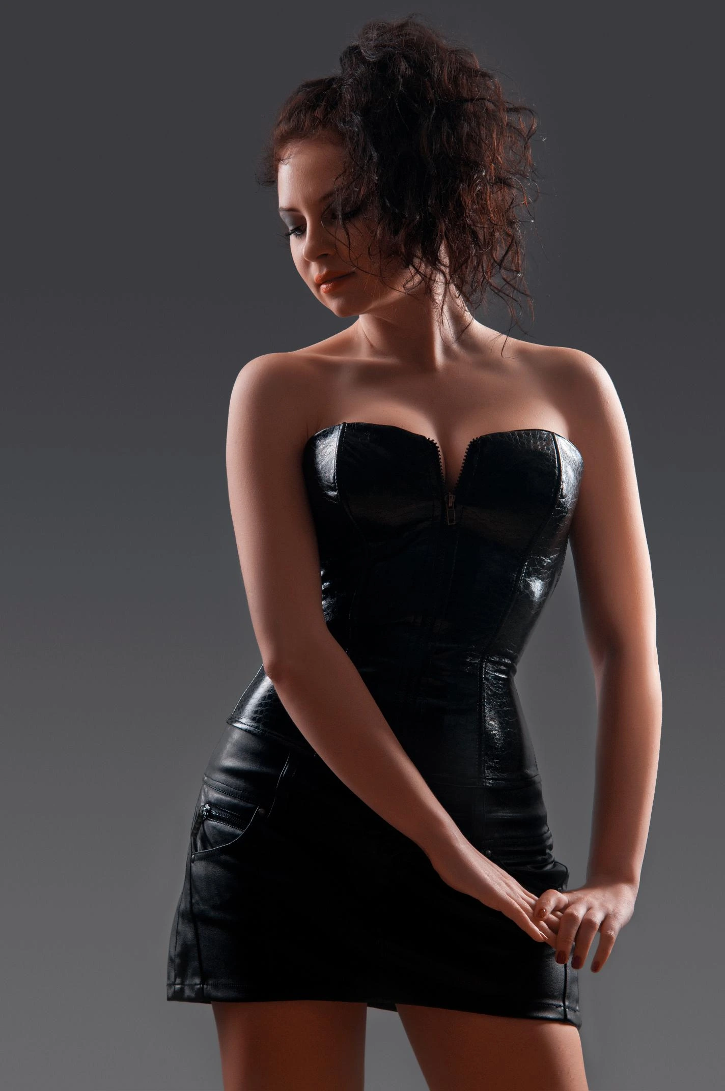 Woman in sleek black PVC mini dress with zipper front posing under studio light.