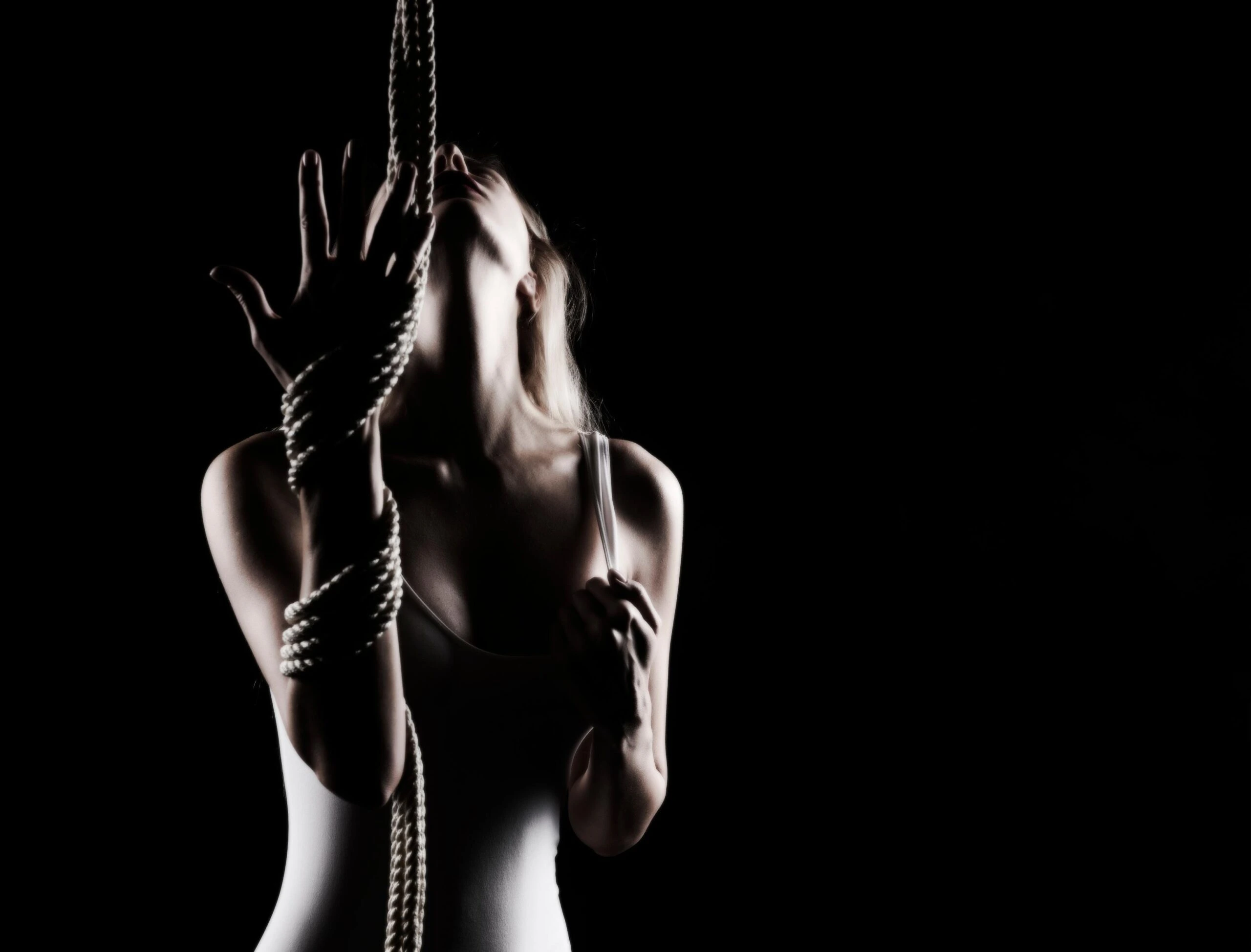 Dramatic shot of woman in white bodysuit bound in rope, reaching toward the ceiling in low light