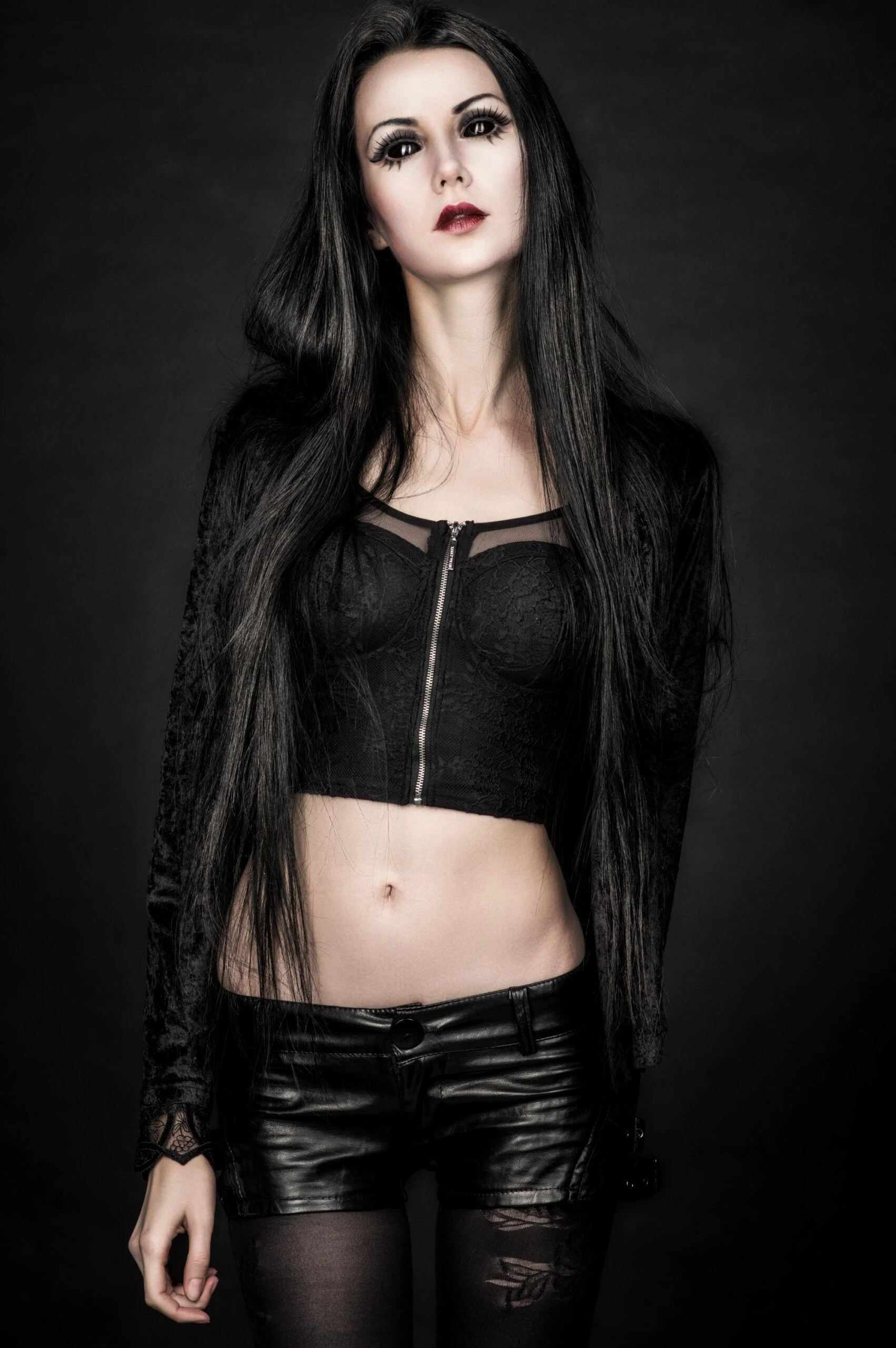 Gothic woman in black lace crop top and leather shorts standing against dark background.