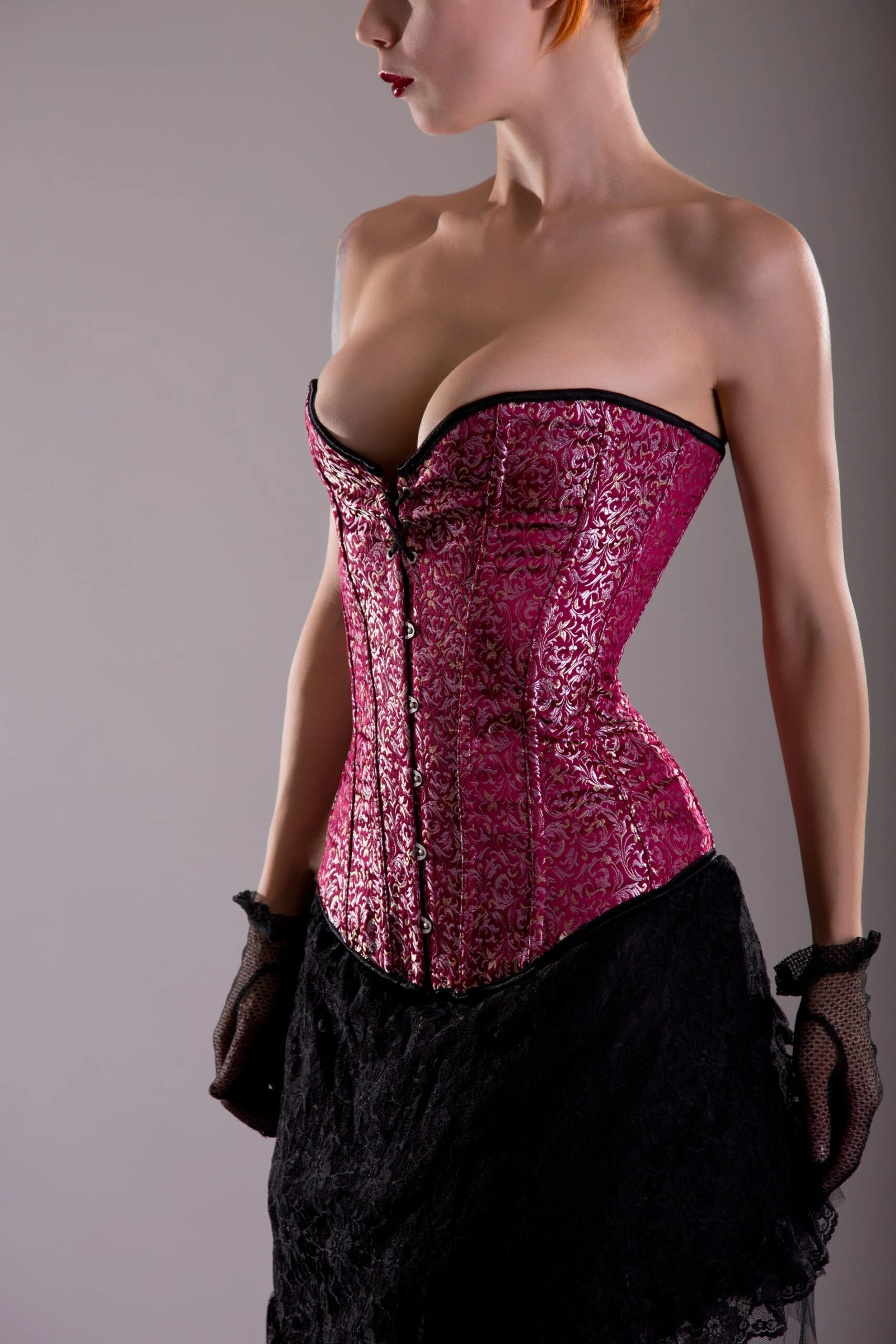 Woman in a pink brocade overbust corset with black lace posing in profile.