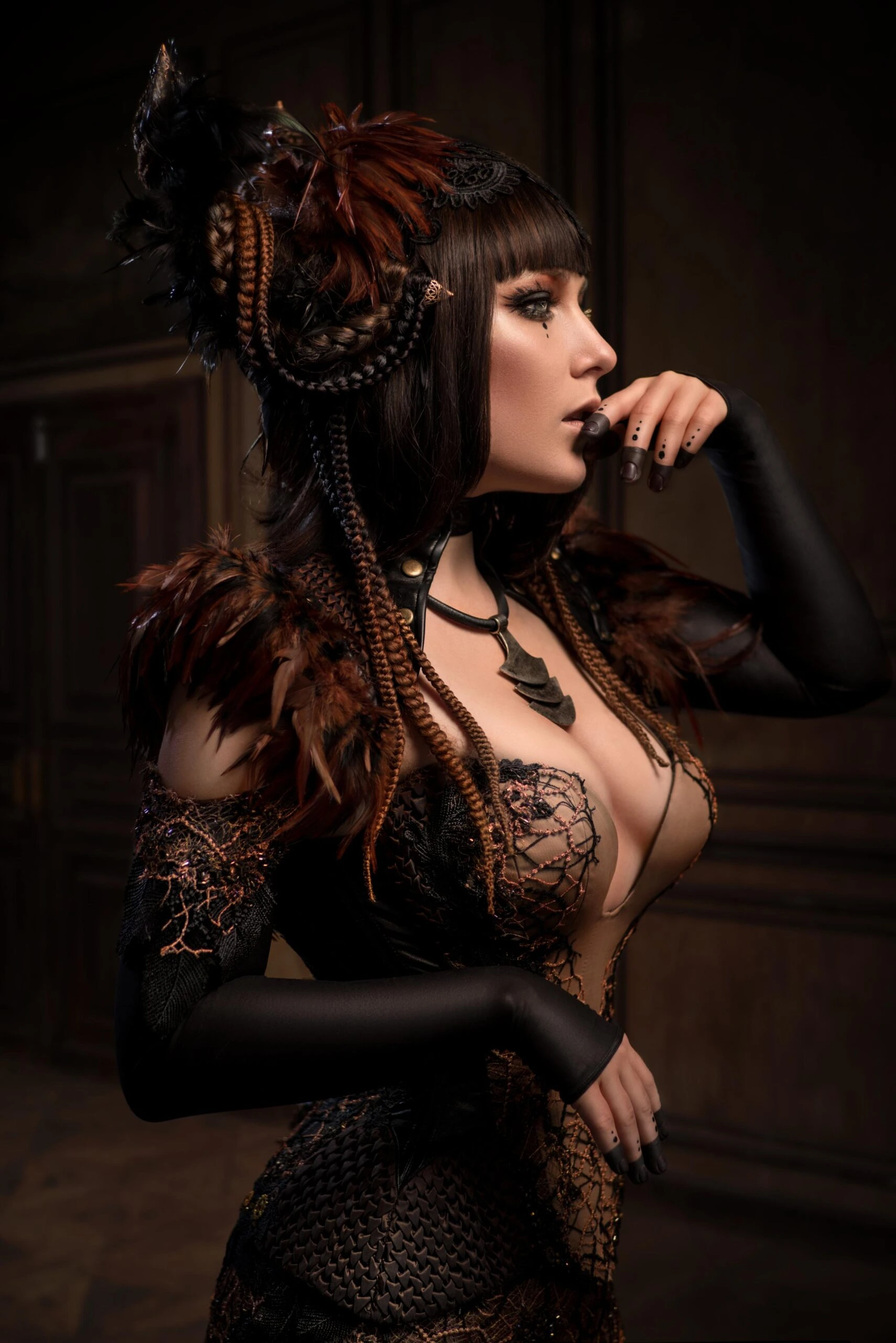 Profile portrait of a woman with braided hair, feather details and dark makeup, wearing an ornate dark outfit in warm lighting.