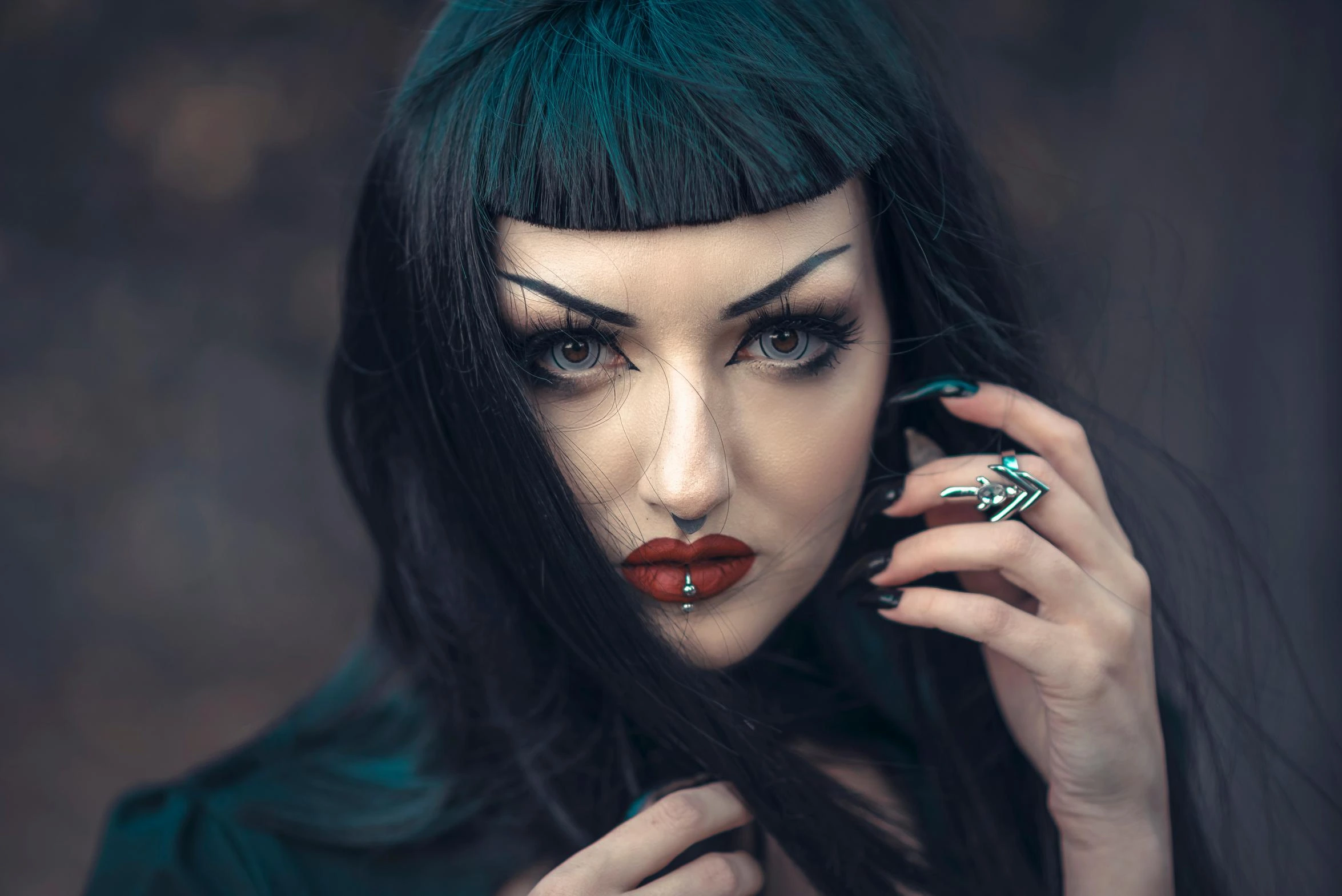 Close-up portrait of a gothic woman with blunt bangs, dark makeup, red lips, facial piercing, and intense gaze against a blurred background.