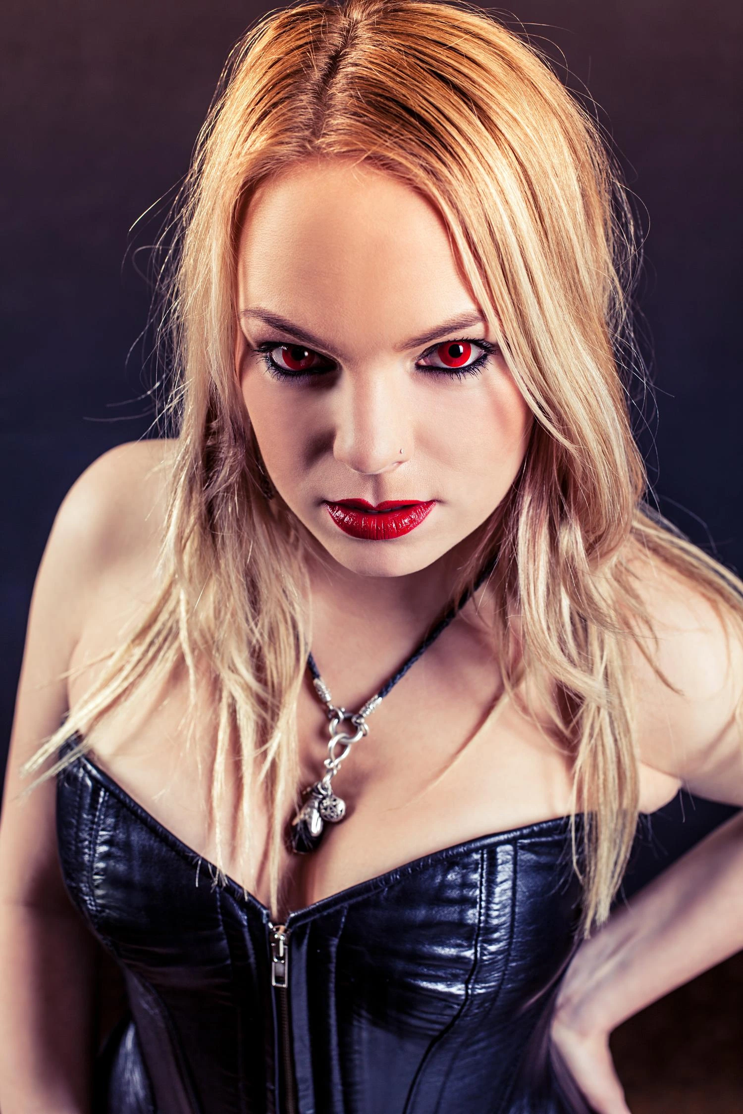 Blonde woman wearing a black corset with red lipstick and red contact lenses, intense close-up portrait against a dark background.