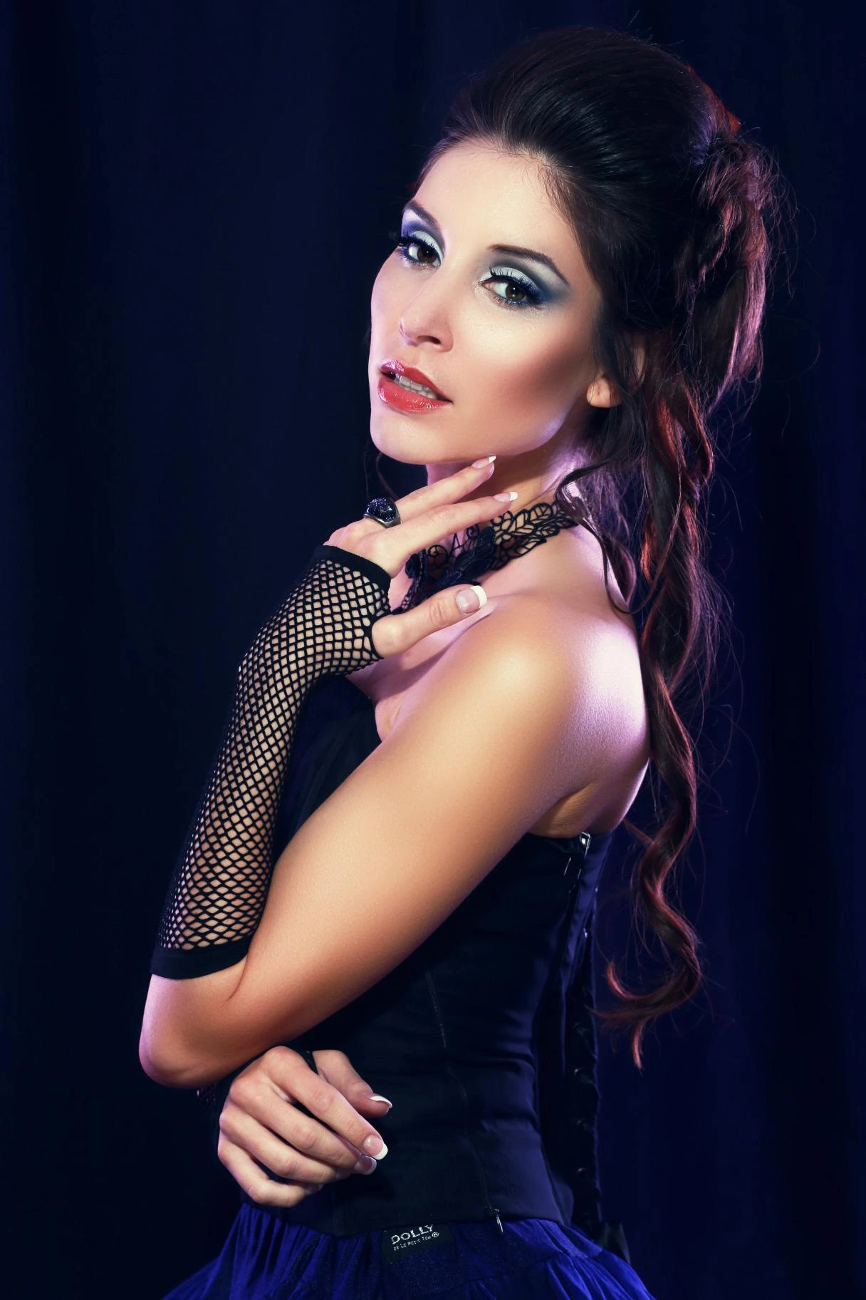 Studio portrait of a woman in a black corset dress with dramatic makeup and lace glove, posing against a dark blue background.