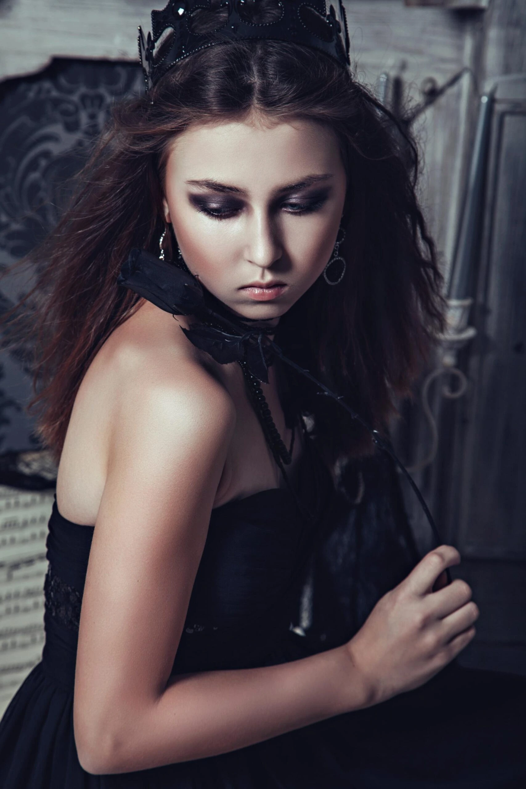 Gothic-style portrait of a woman wearing a dark crown and black dress, looking down with moody makeup in a vintage interior setting.