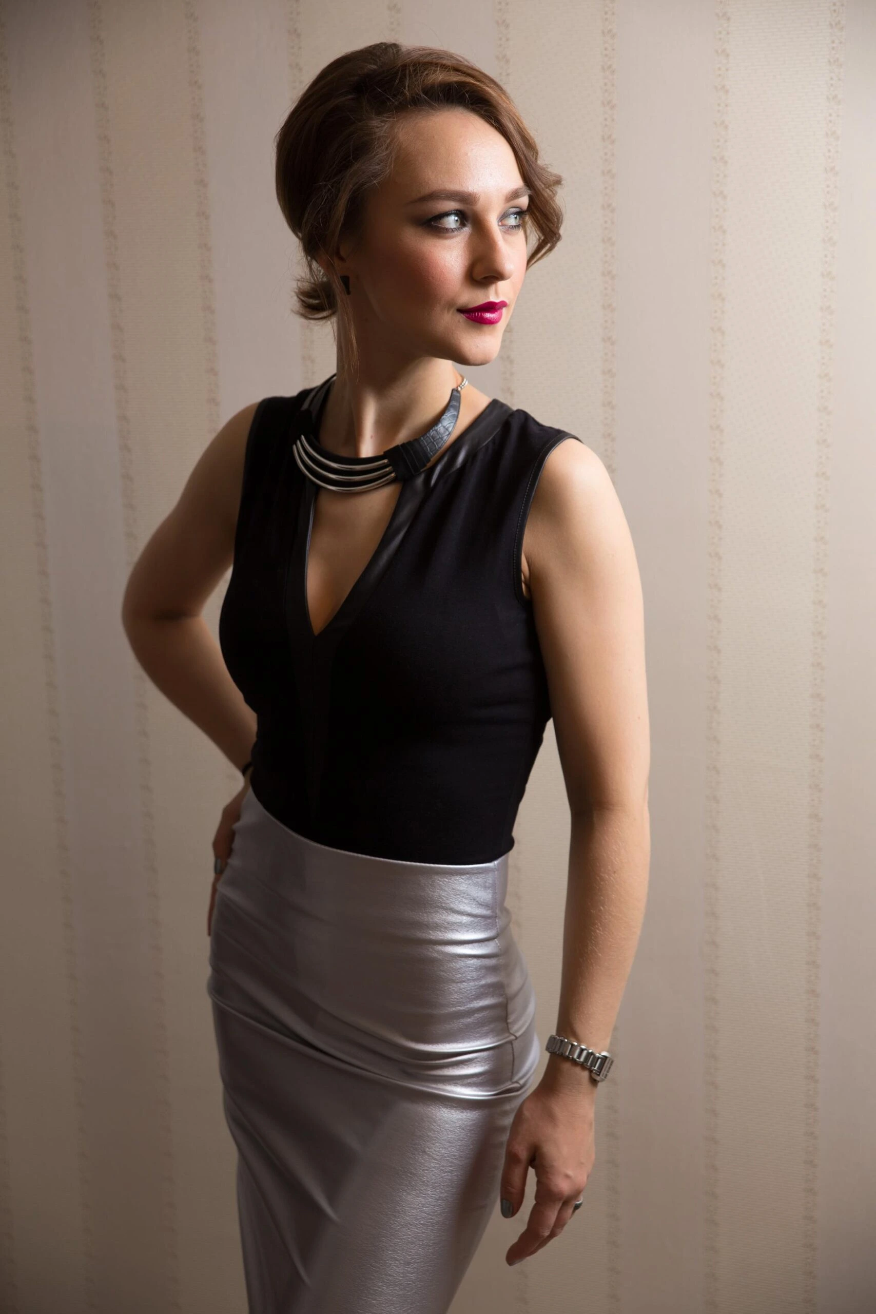 Elegant woman in a black sleeveless top and metallic silver skirt standing indoors, softly lit portrait with neutral background.