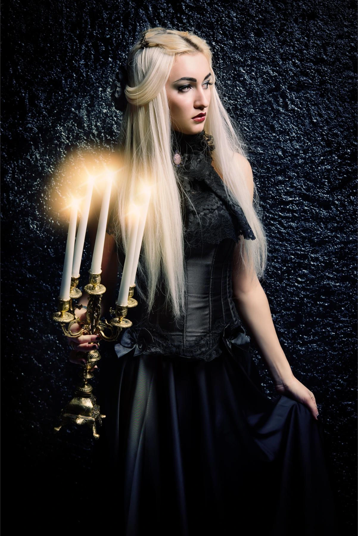 Blonde woman in a dark gothic dress holding a brass candelabra with lit candles against a textured black wall.
