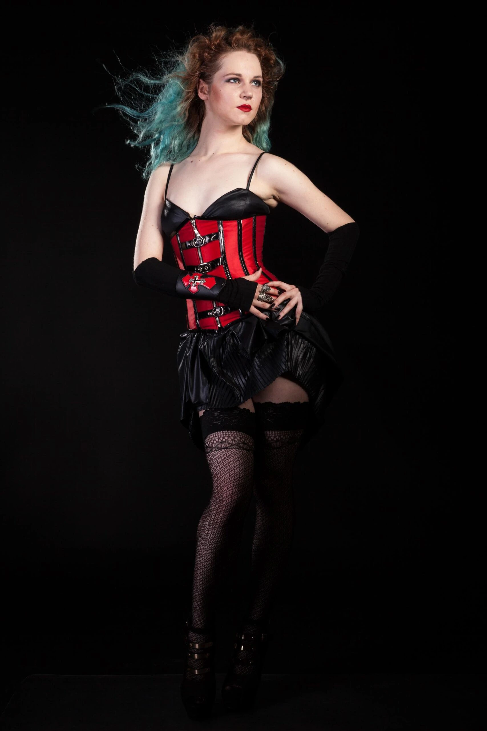 Woman in a red and black corset with black skirt and fishnet stockings, posing confidently against a dark studio background.
