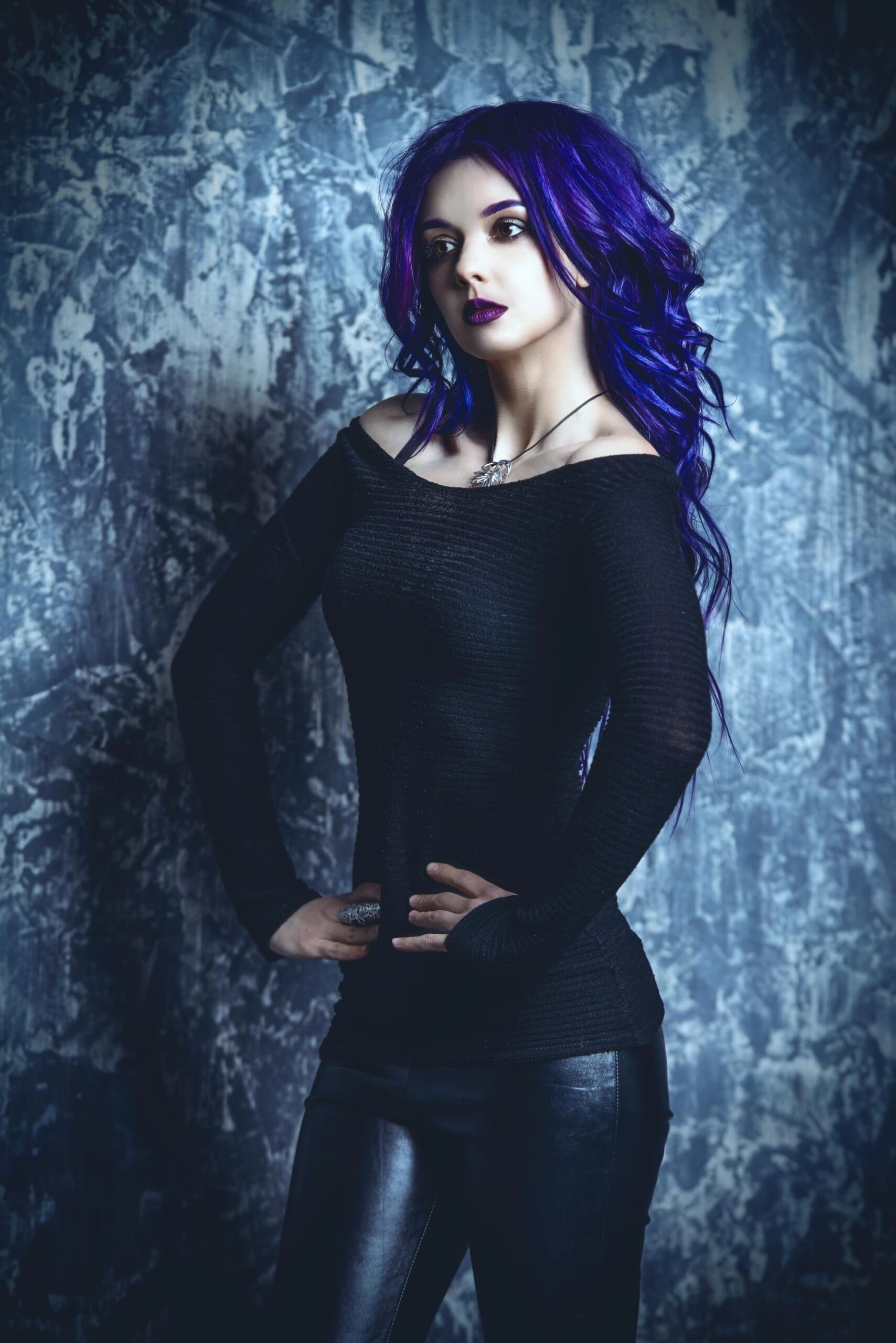 Woman with purple hair wearing a fitted black top, hands on hips, standing against a textured blue-gray wall.