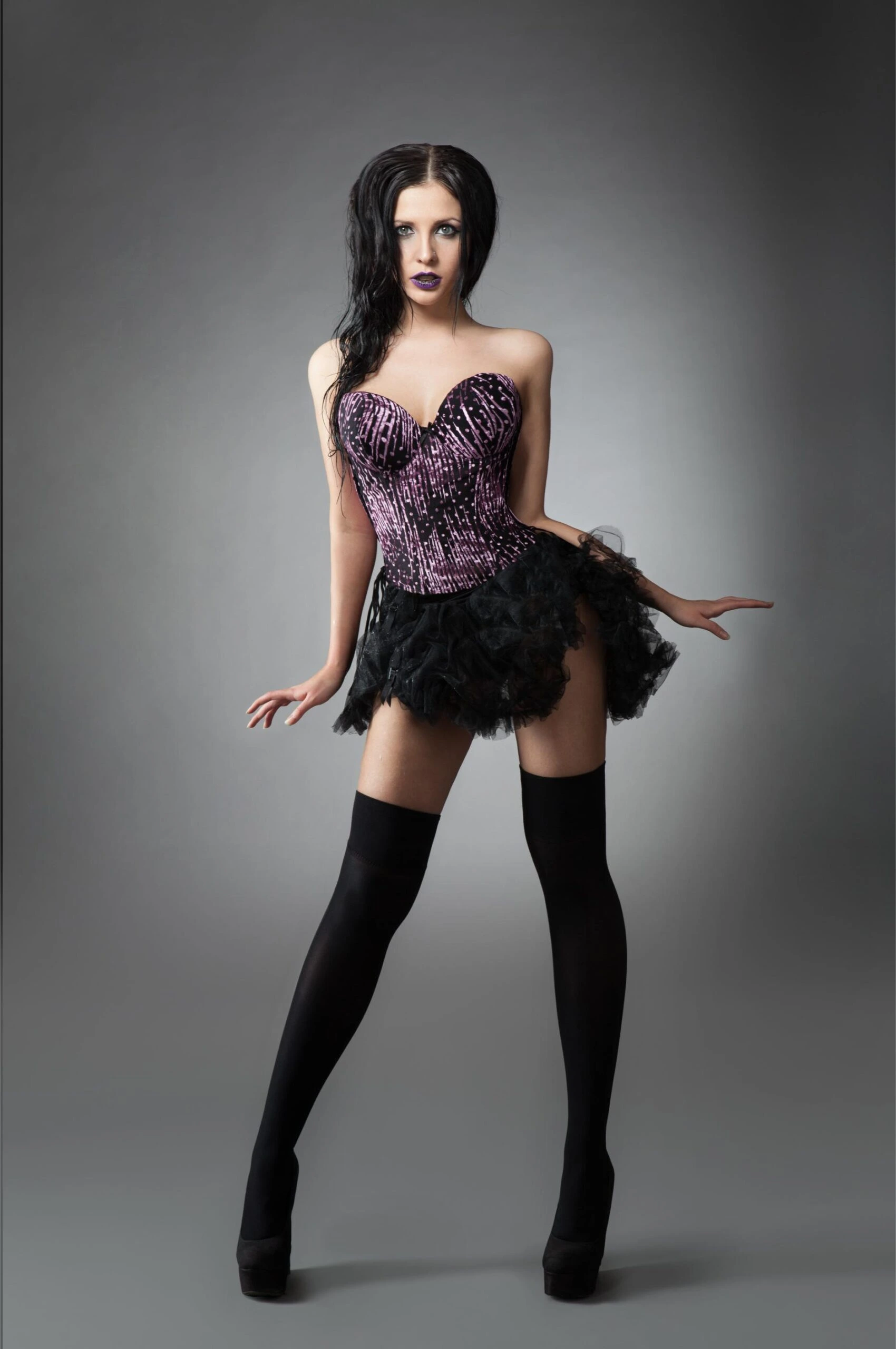 Woman in purple corset, short black skirt and thigh-high stockings posing against gray studio background.