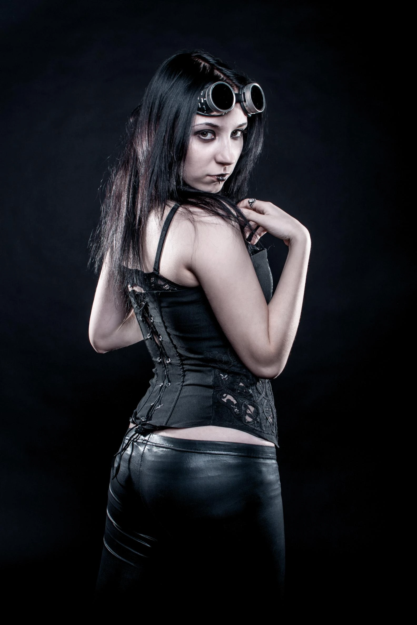 Woman in black corset and leather pants with steampunk goggles, posing over shoulder on dark background.