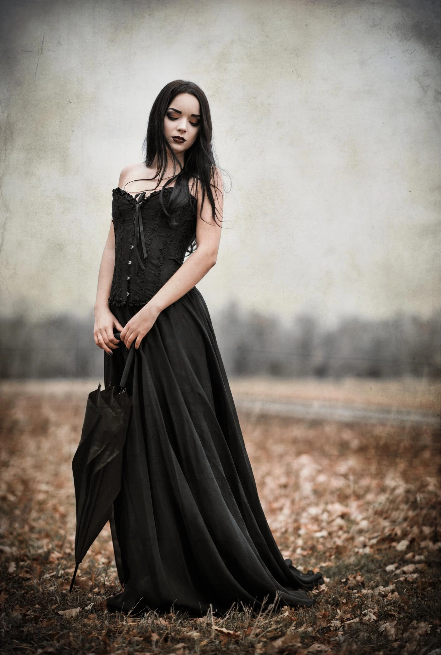 Woman in long black corset dress holding a closed umbrella, standing in a leaf-covered field under an overcast sky.