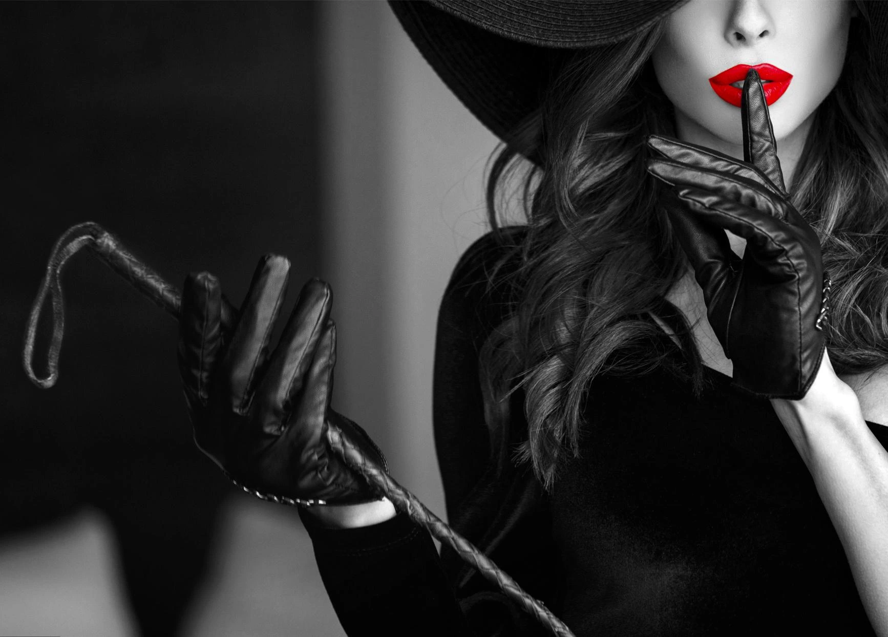 Black-and-white portrait of a woman with red lipstick and gloves, holding a whip and making a quiet gesture.